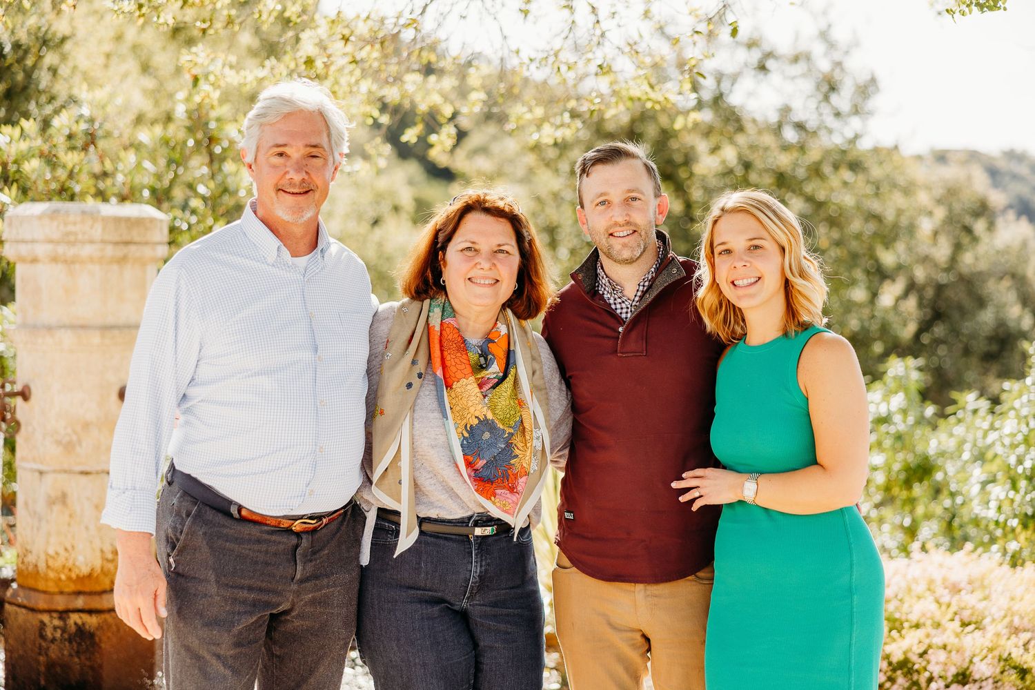 Family group posing together outdoors in warm sunlight with trees and greenery in the background.