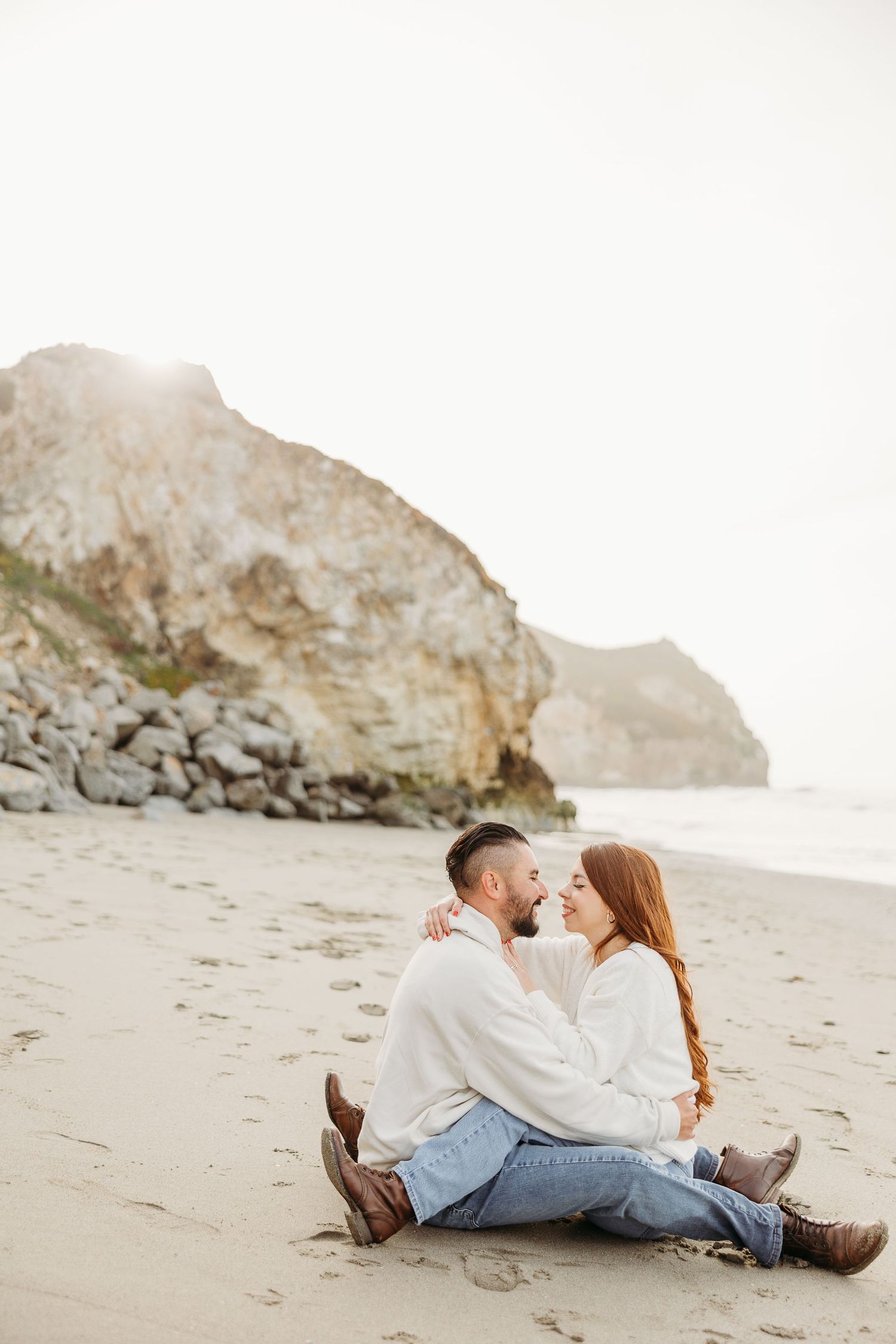 A couple sits close together on a sandy beach with dramatic coastal cliffs and misty shoreline in the background.
