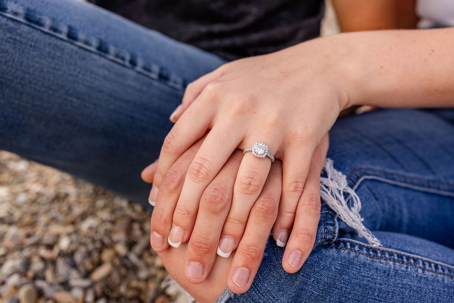 Close-up of hands showing an engagement ring while sitting on a pebbly beach.