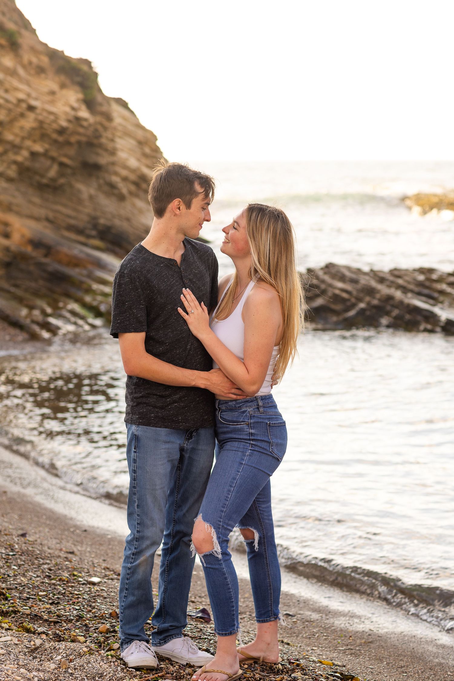 A couple shares a tender moment on a beach with rocky cliffs in the background.