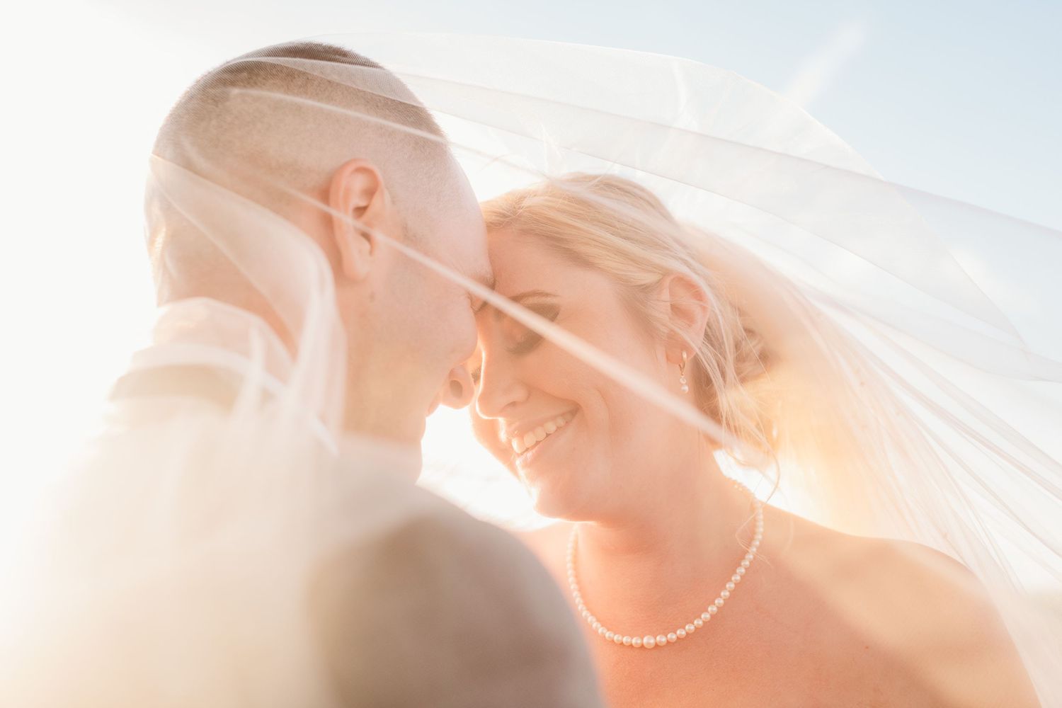 A close-up wedding photo showing a couple's intimate moment under a sheer veil with sunlight shining through.