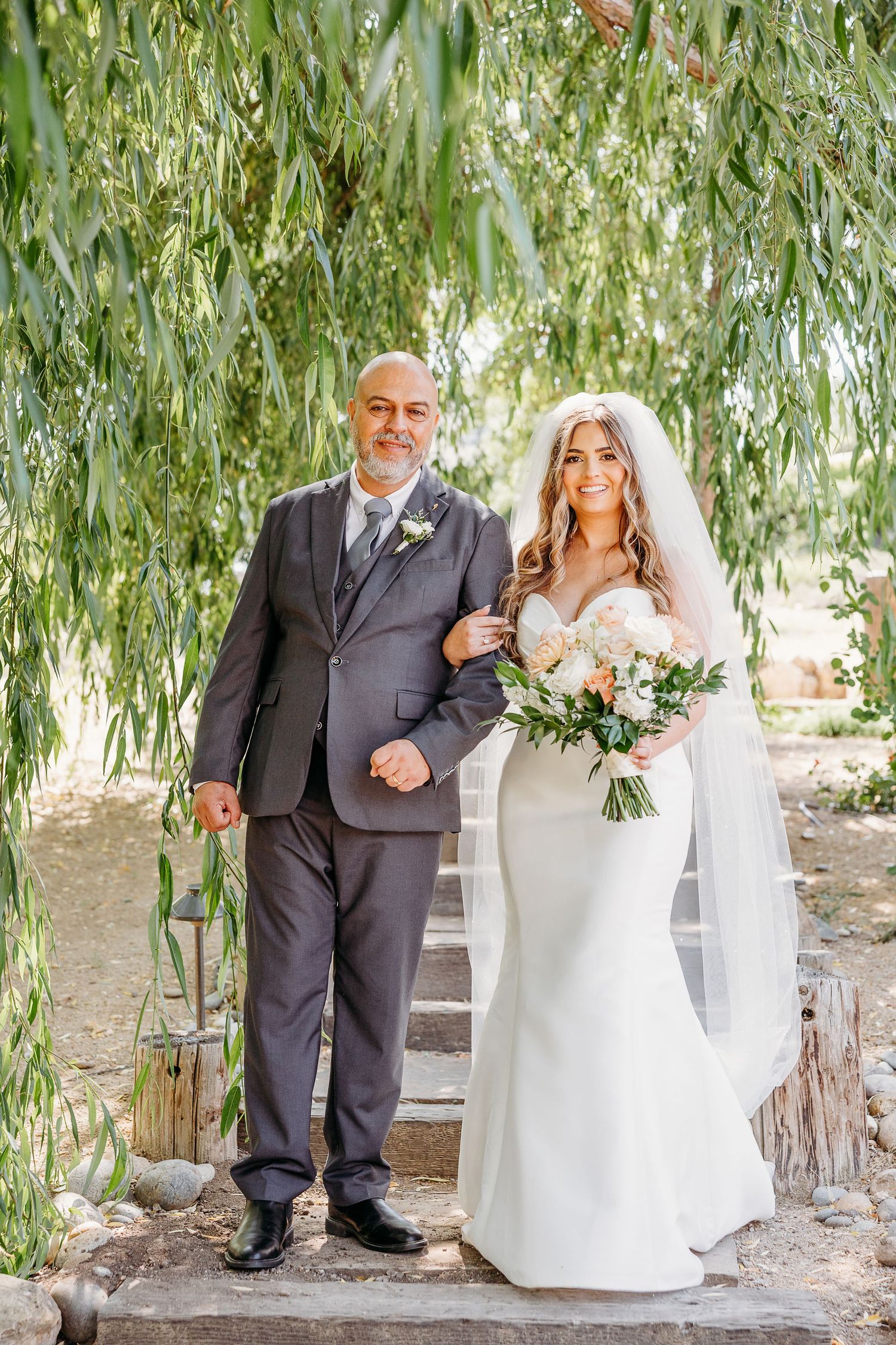 An elegant wedding portrait under a willow tree with cascading branches and natural stone steps.