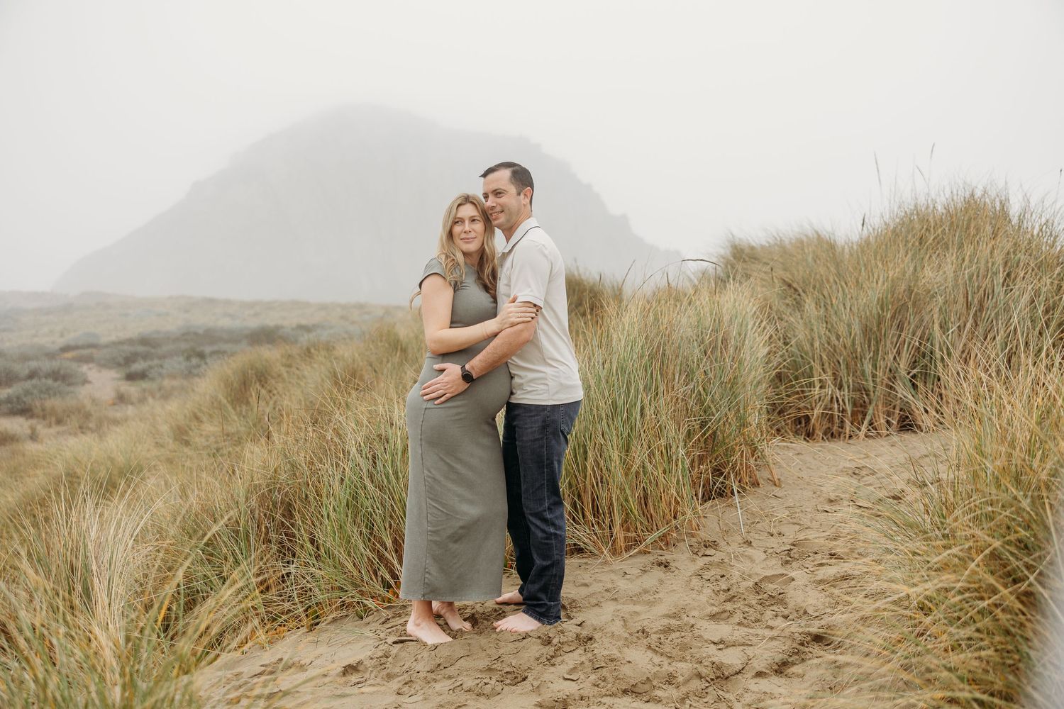 A romantic maternity photo captures a couple standing together among coastal dunes at sunset.