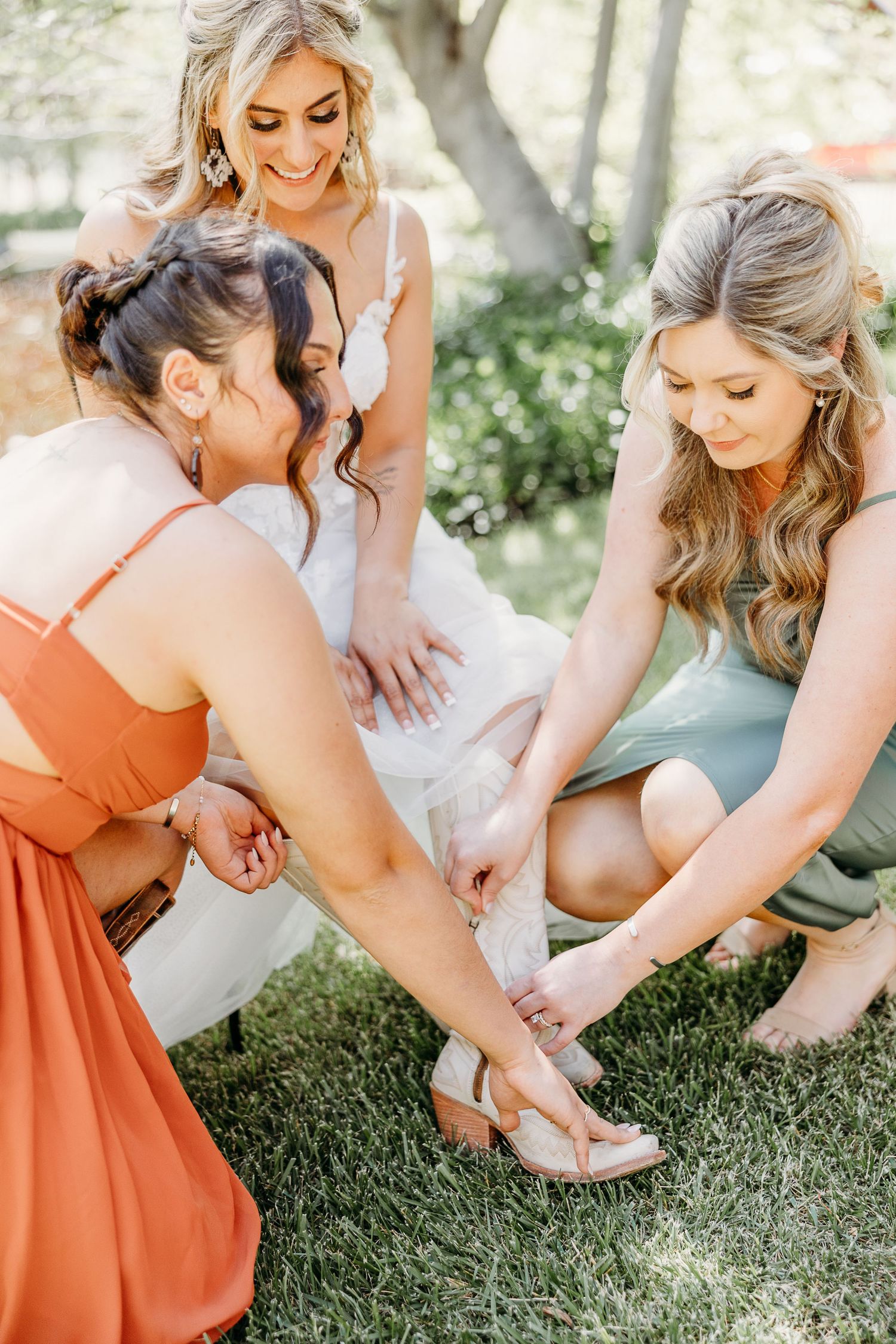 Bridesmaids in coral dresses help adjust the bride's shoes on a grassy lawn.
