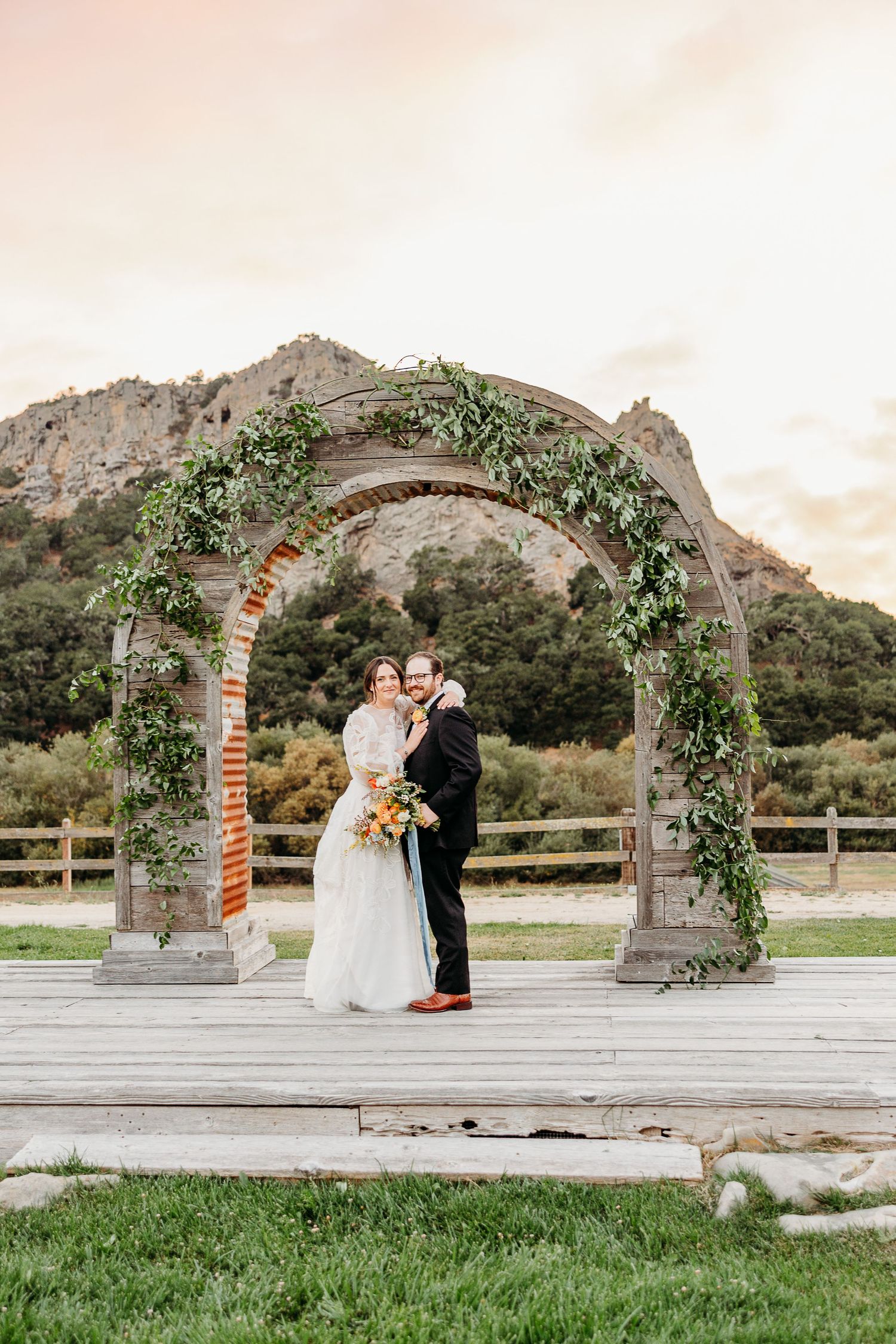 Couple embracing under stone archway decorated with climbing vines at outdoor wedding venue by leading San Luis Obispo wedding photographer.