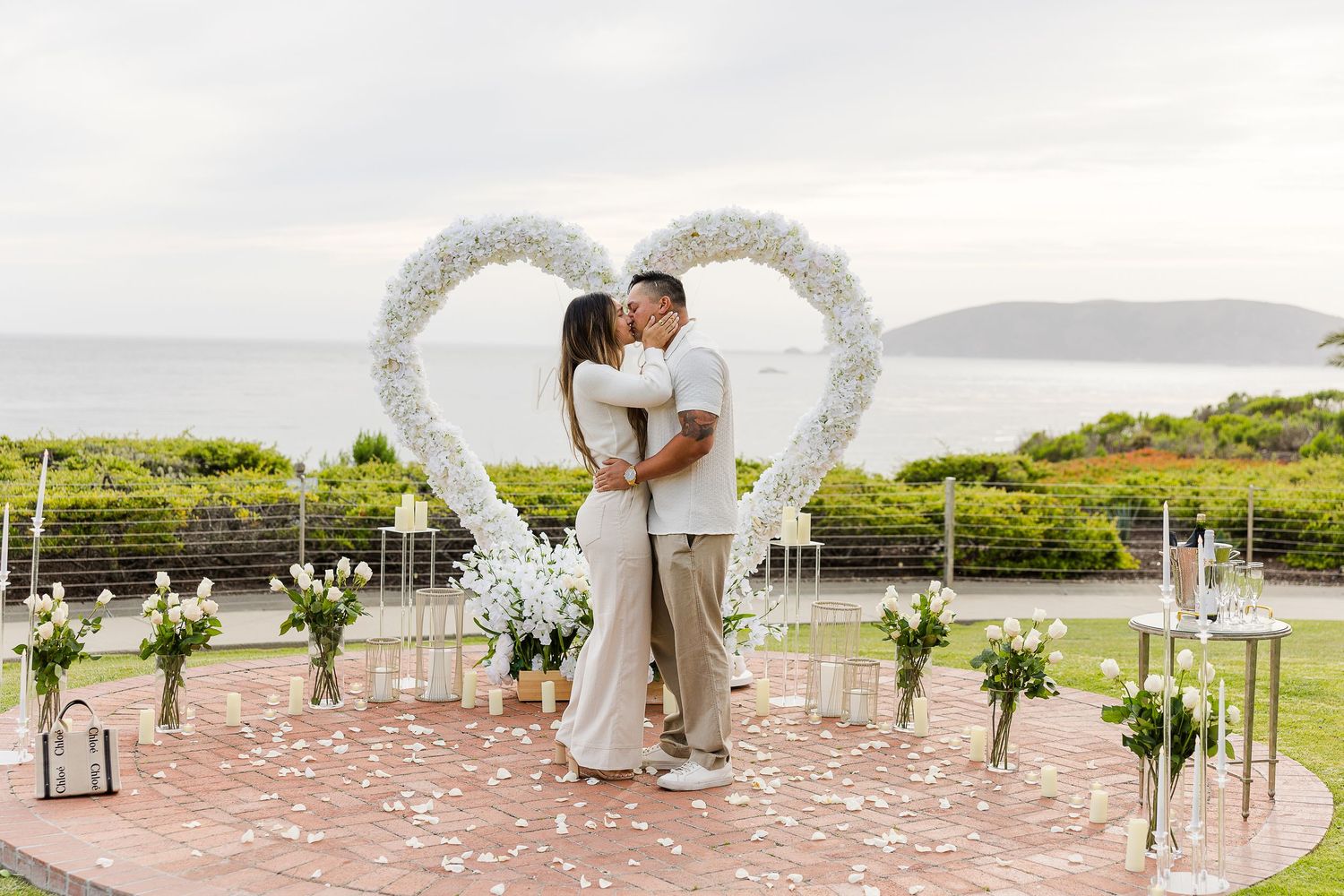 A romantic outdoor wedding ceremony setup with white chairs and floral arrangements on a brick patio overlooking a scenic view.