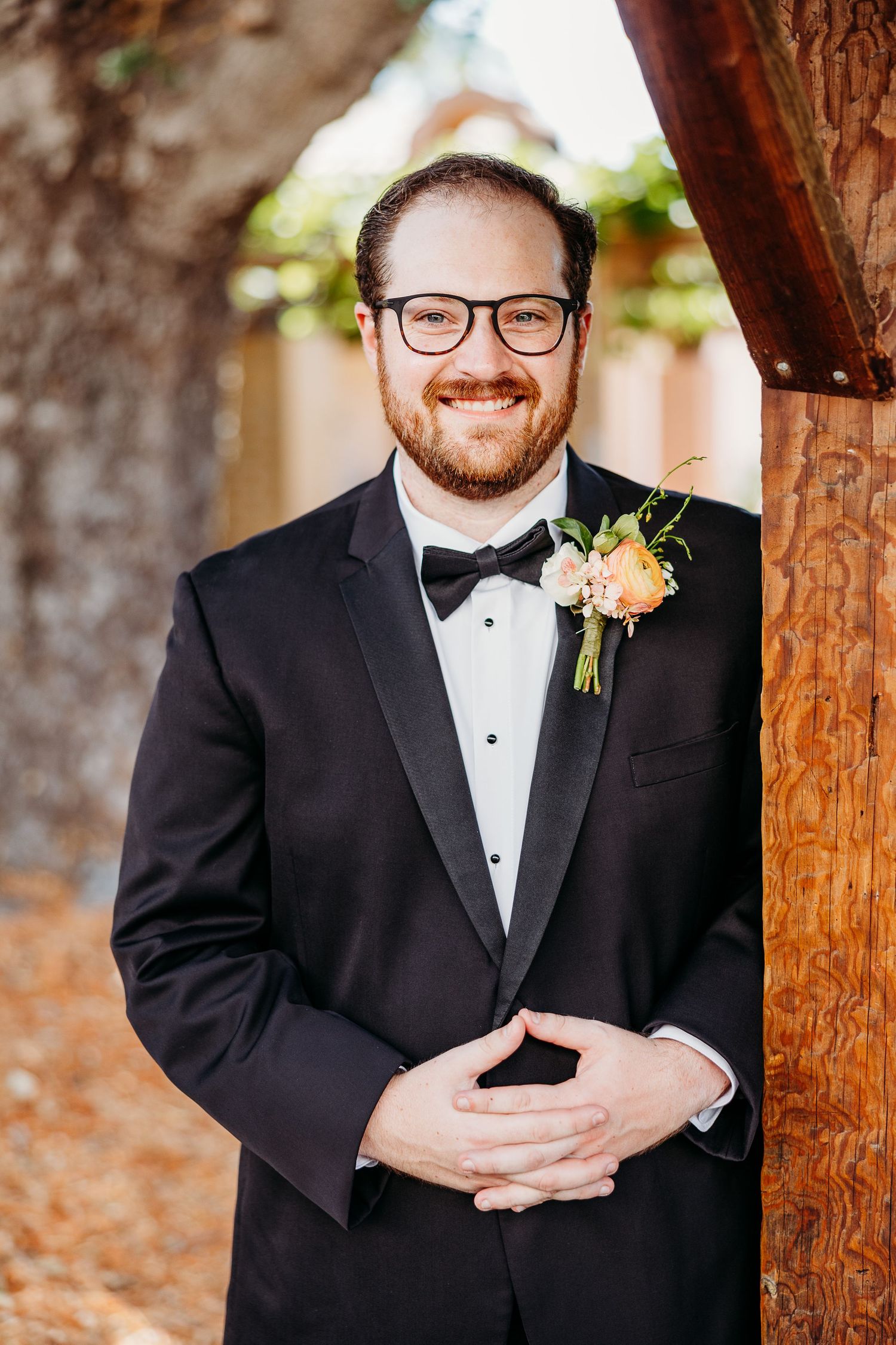 Groom in formal tuxedo with boutonniere smiles warmly while standing by wooden post.