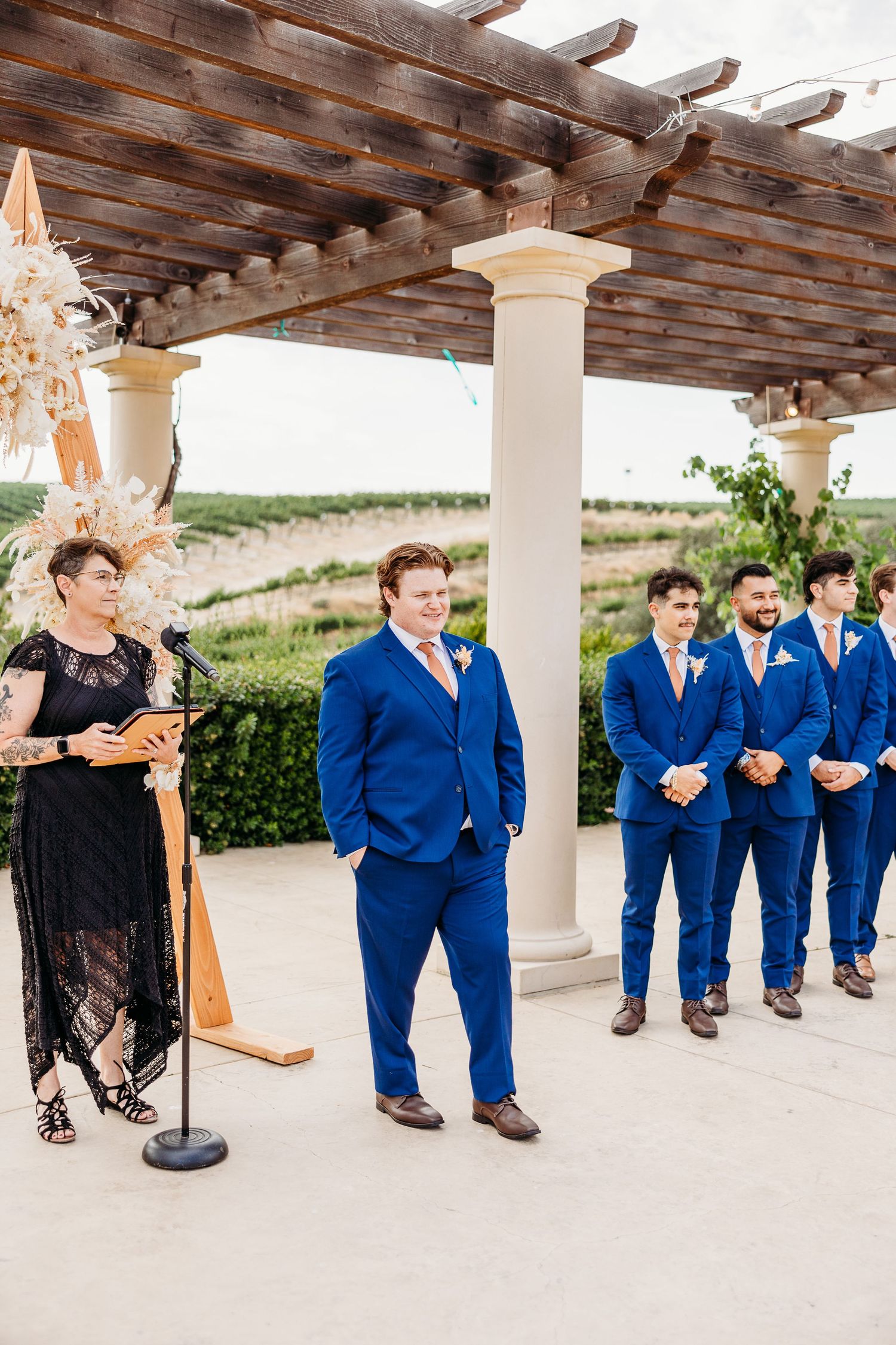 Groomsmen in bright blue suits stand under a wooden pergola at an outdoor wedding venue.