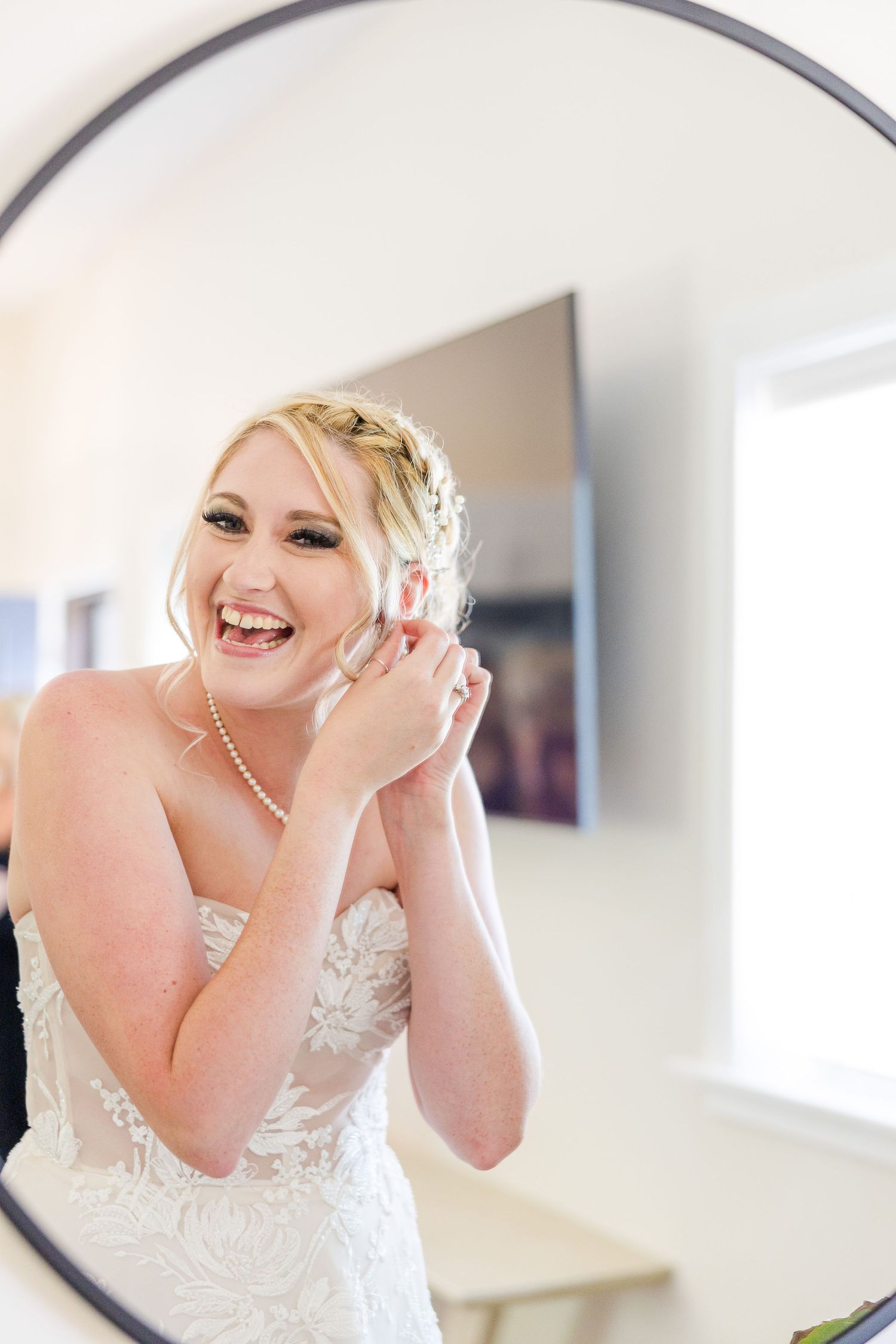 Mirror reflection captures radiant moment of bride wearing pearl necklace and lace wedding gown.