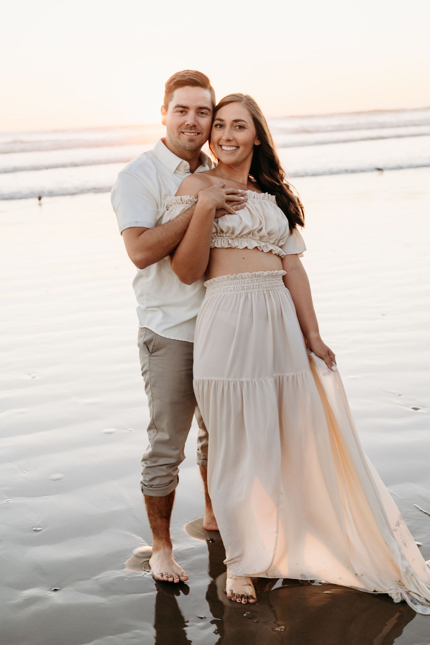 Couple poses together on beach at sunset in coordinated white and beige outfits.