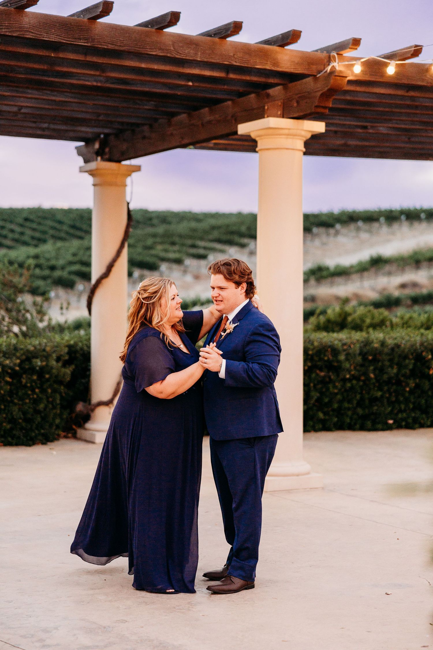 A couple in navy blue outfits dance beneath a wooden pergola at sunset.