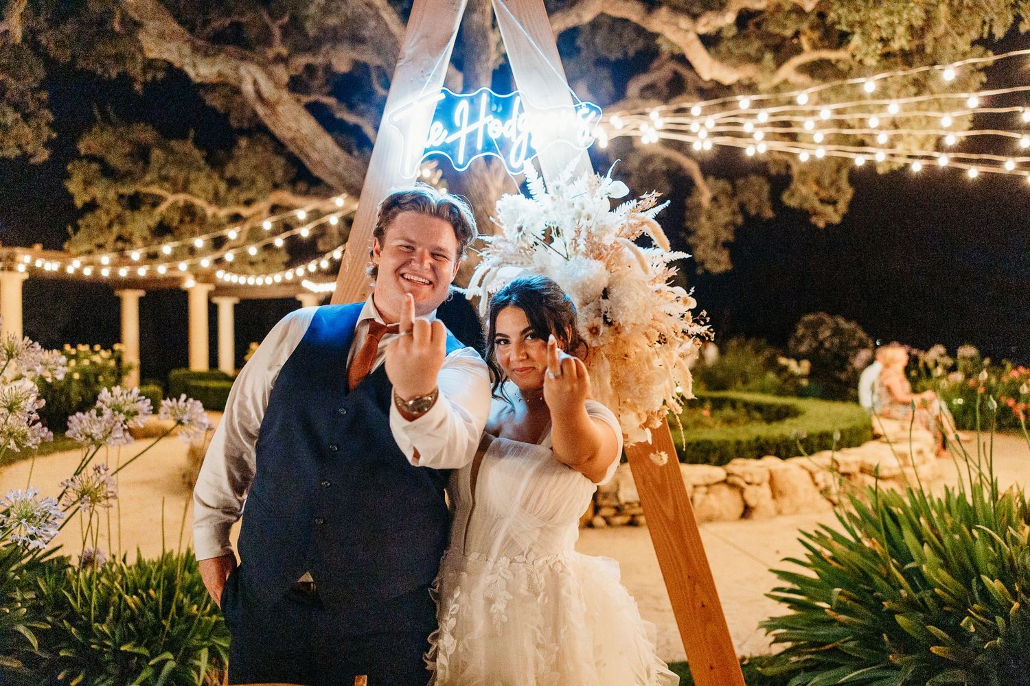 Happy couple posing playfully under triangle arch with neon lighting at evening reception.
