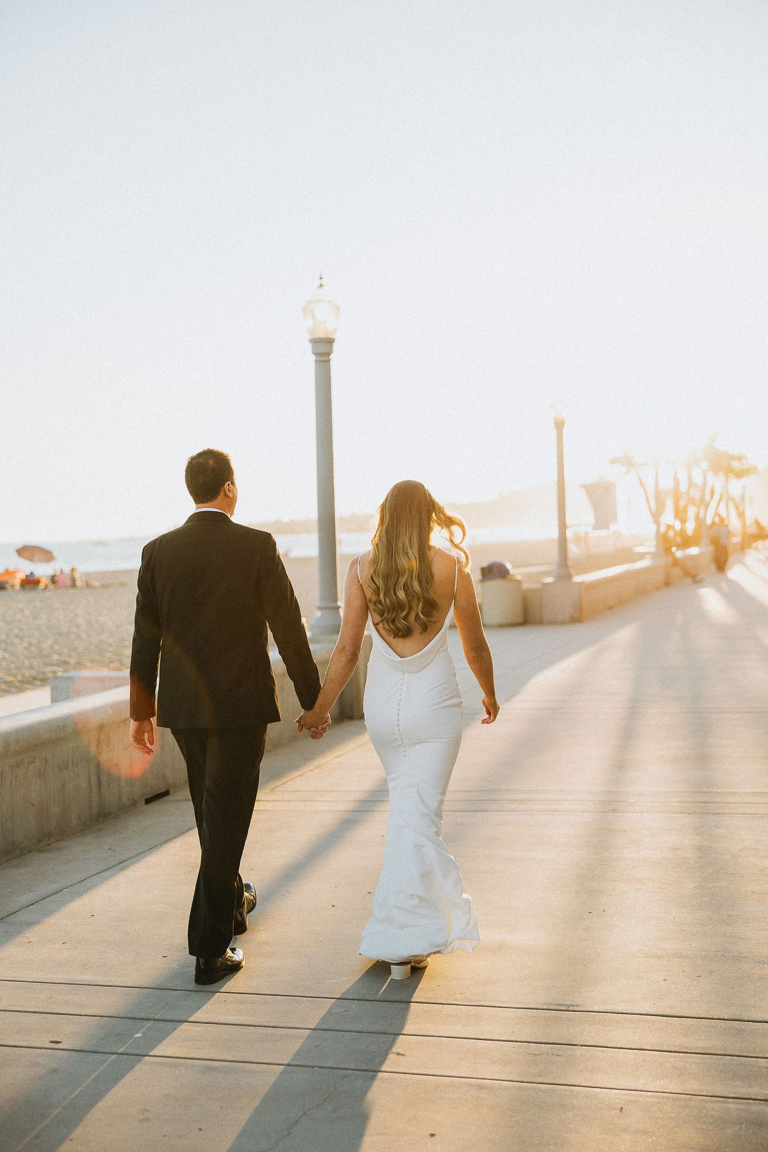 A couple walks hand in hand along a sunset boardwalk by the ocean.