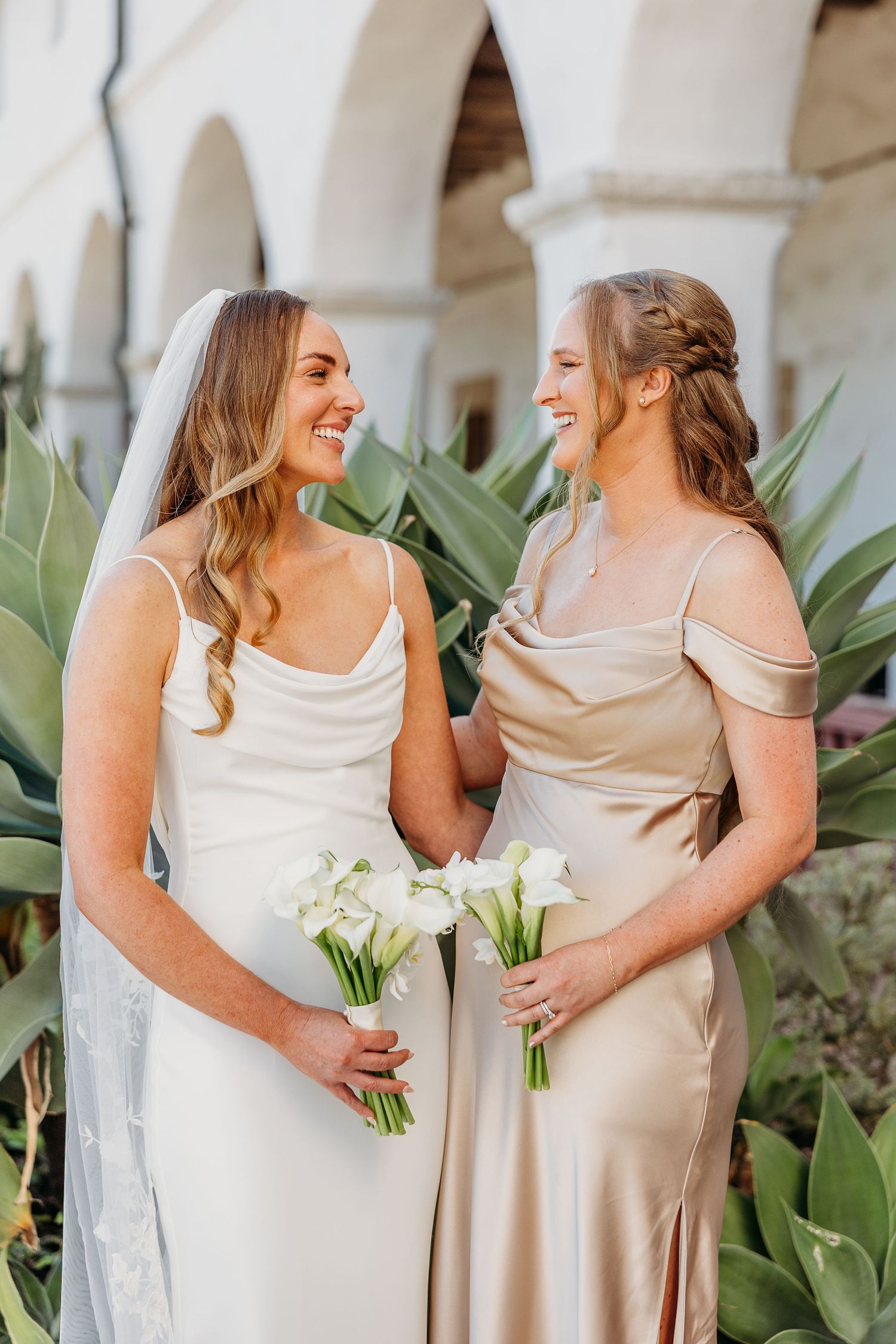 Two women in elegant dresses share a joyful moment together against a backdrop of tropical plants.