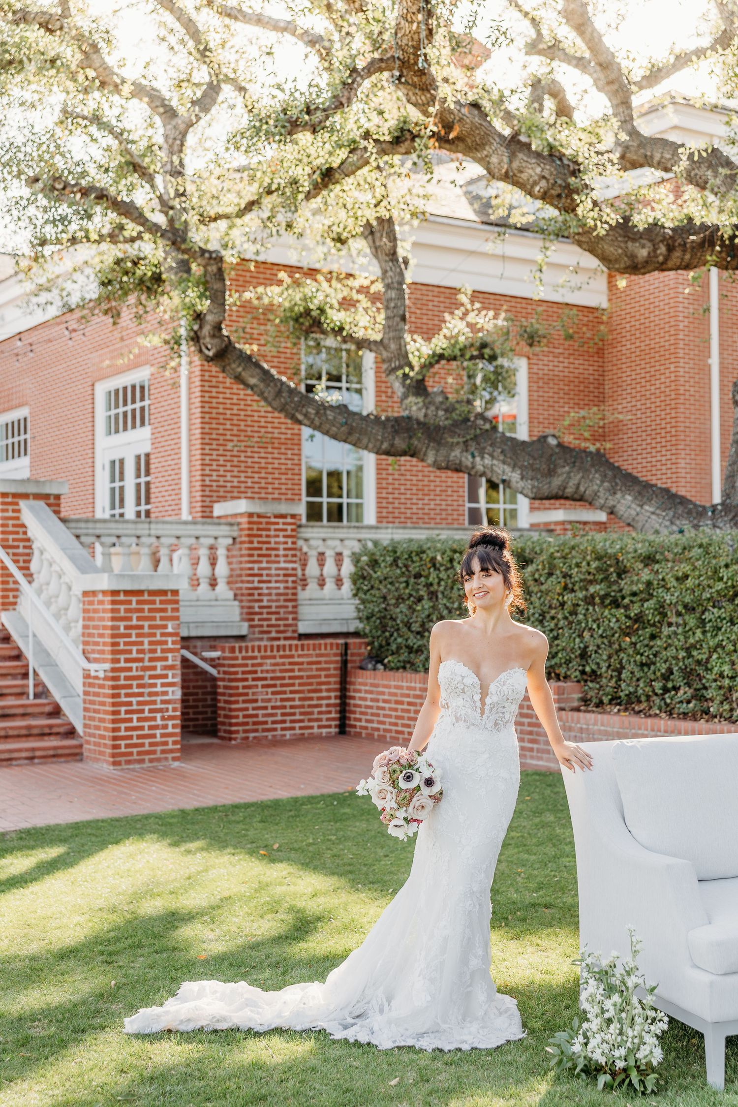 Elegant white wedding gown with lace details photographed in front of a brick building with overhanging trees.