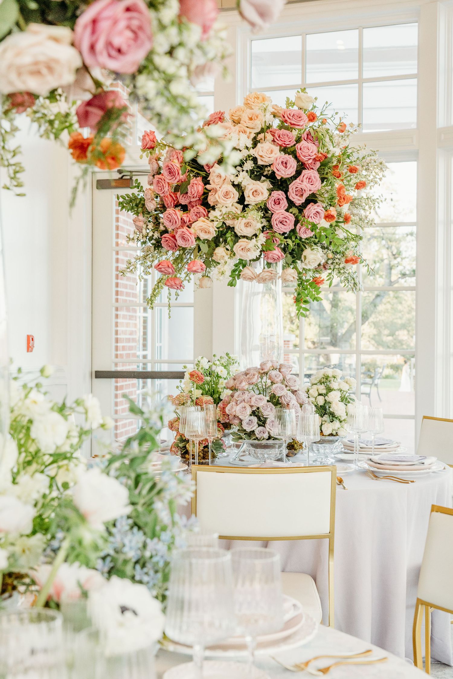 Elegant wedding reception table setting with hanging floral arrangements of pink roses and white flowers in a bright sunlit room.