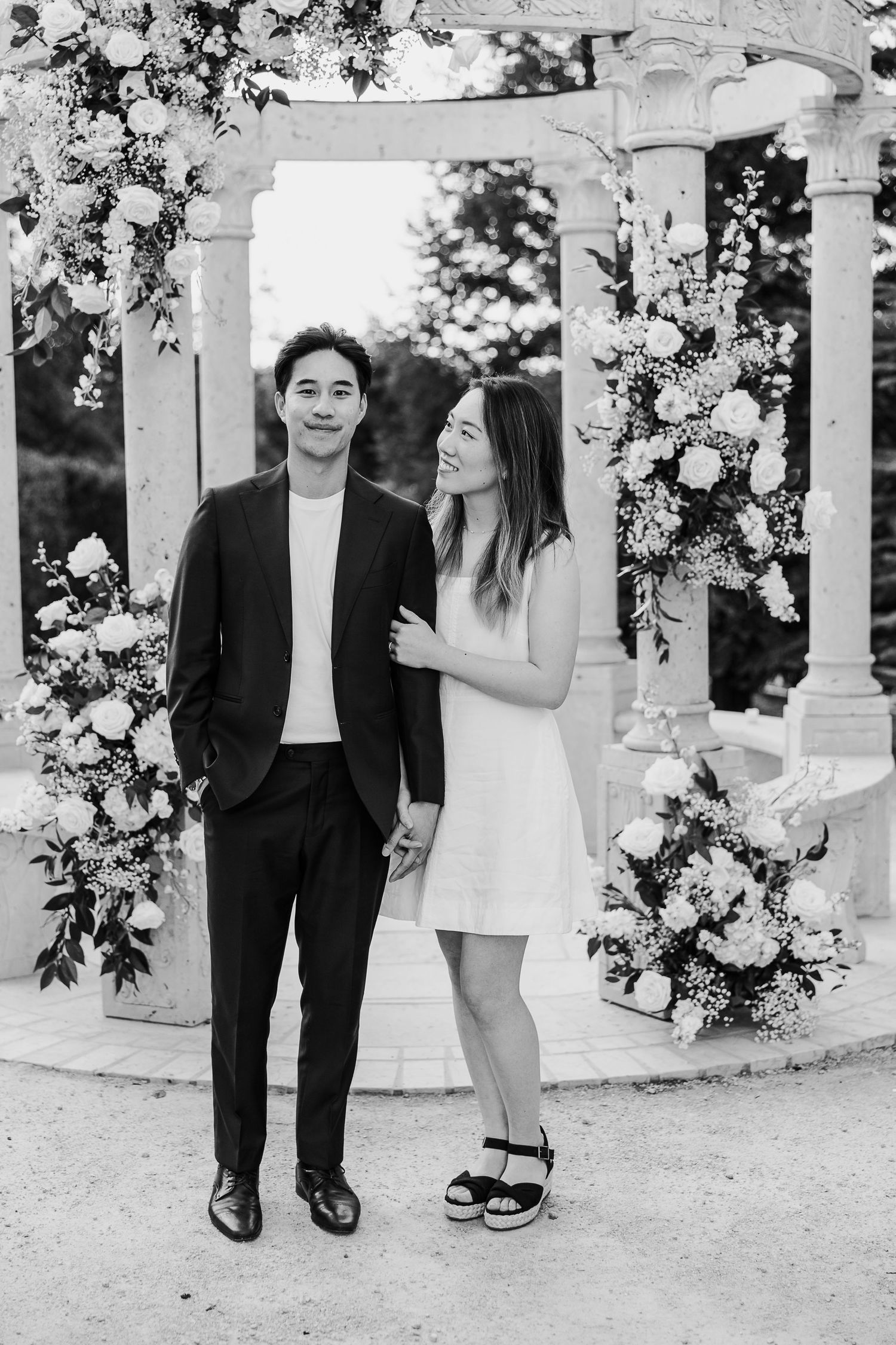 A black and white photo of a couple standing beneath a floral-decorated stone gazebo.