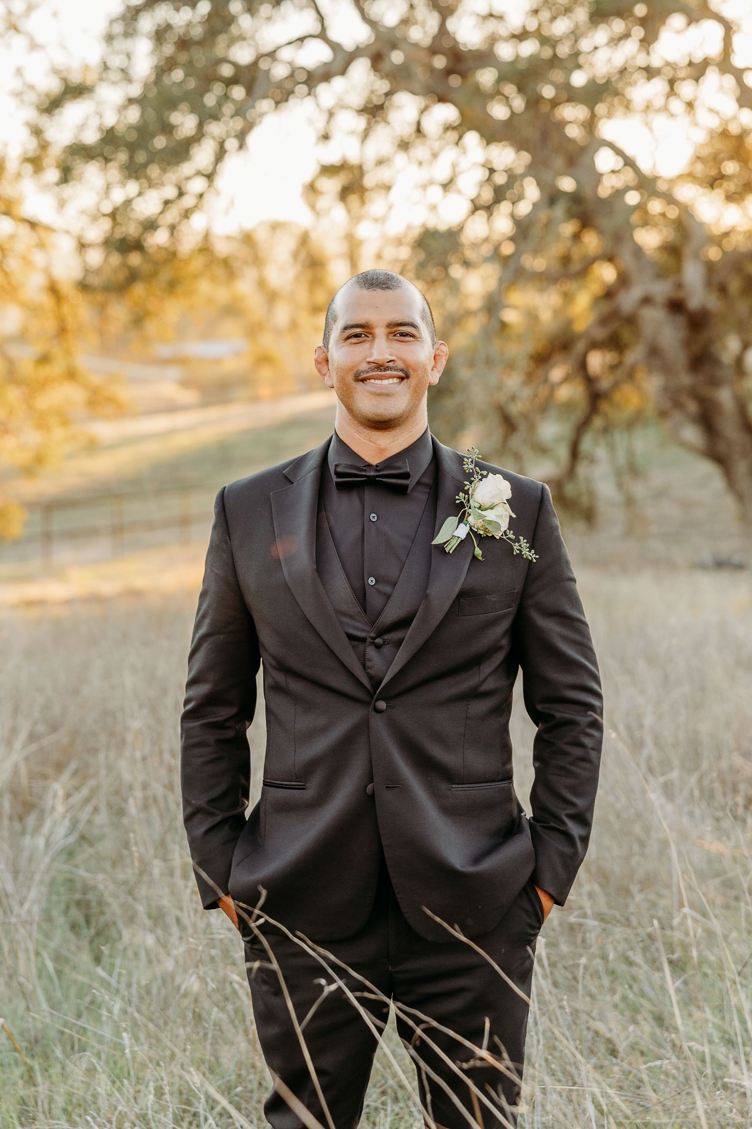 Formal wedding portraits of a groom in a black tuxedo standing in a golden field at sunset.