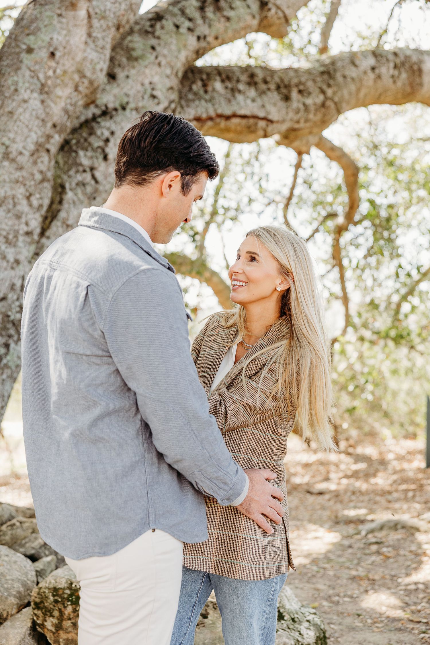A romantic couple embraces outdoors in a natural setting with trees and filtered sunlight in the background.