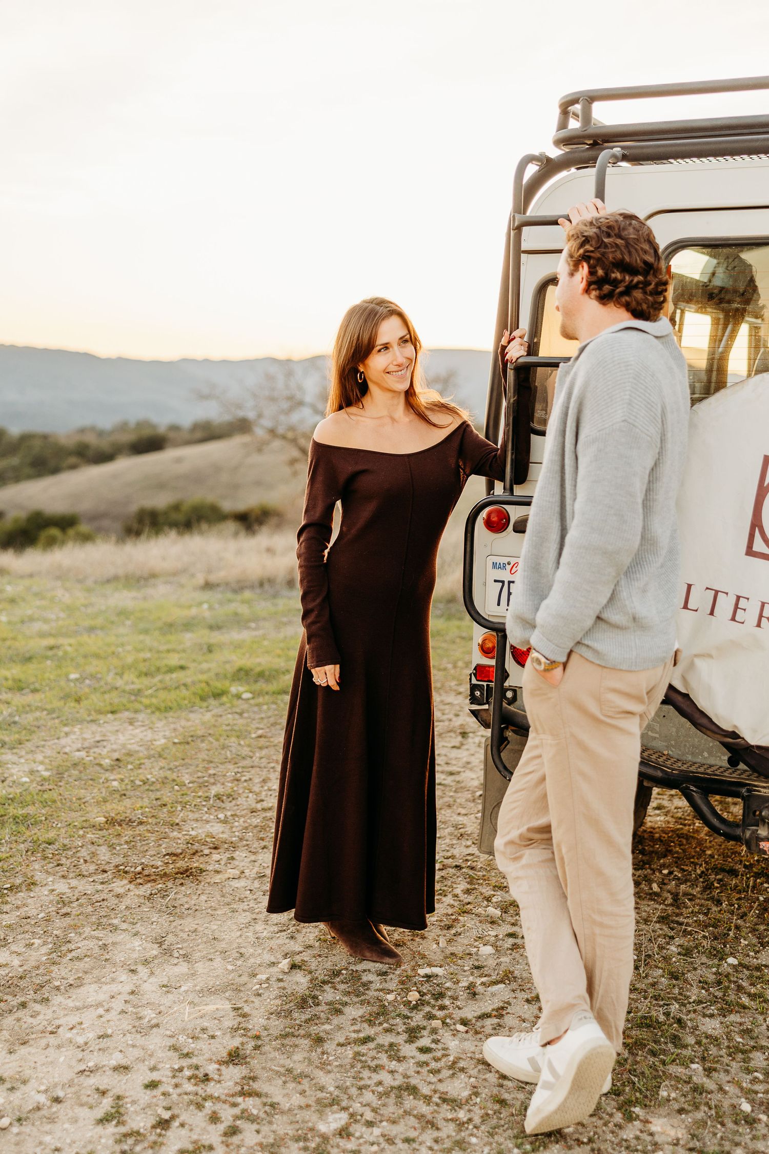 A couple stands near a vintage vehicle on a grassy hillside at sunset, creating a romantic outdoor scene.
