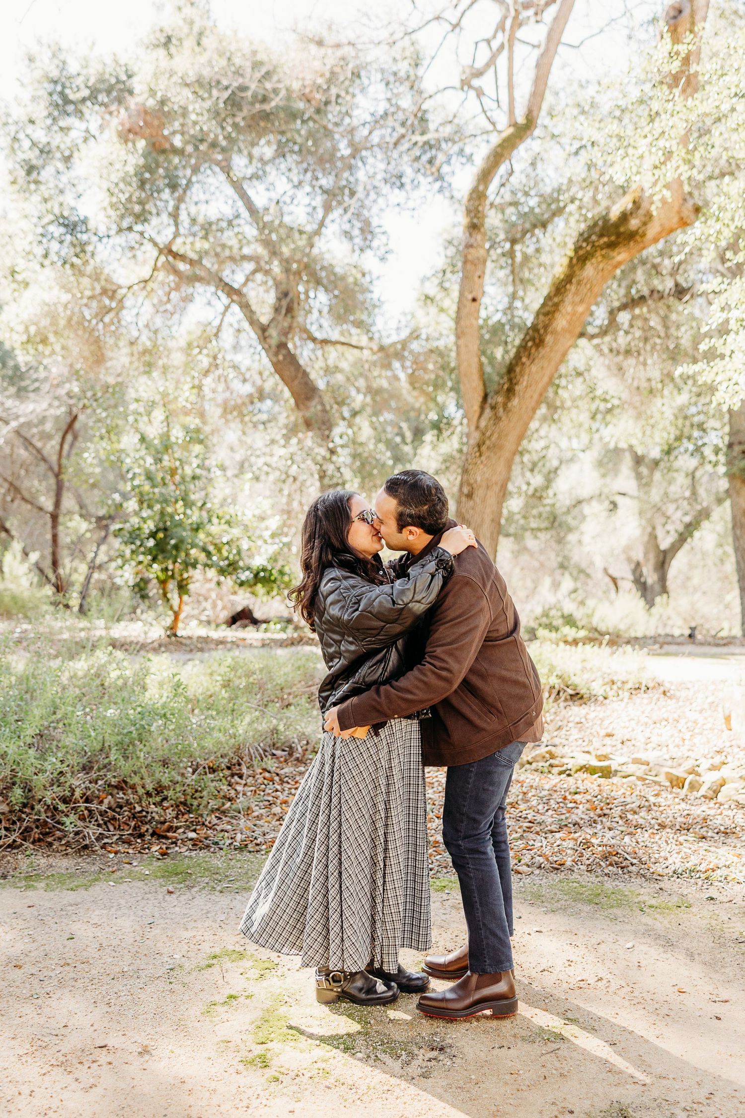 A romantic couple embraces on a path surrounded by olive trees on a sunny afternoon.