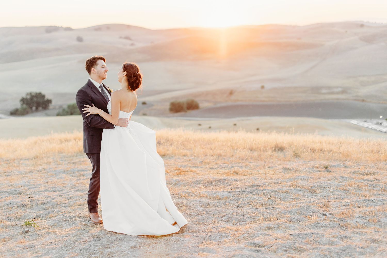 A couple shares an intimate moment against a golden sunset on rolling hills during their wedding photoshoot.