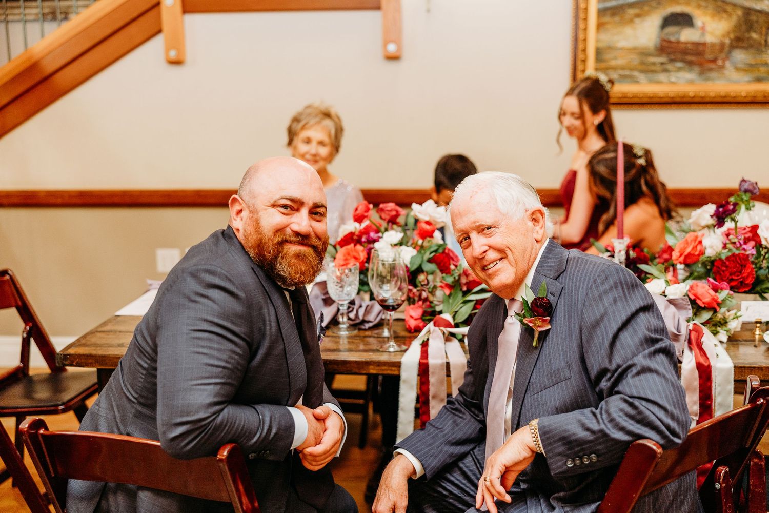 Wedding reception scene with guests seated at tables decorated with red floral arrangements.