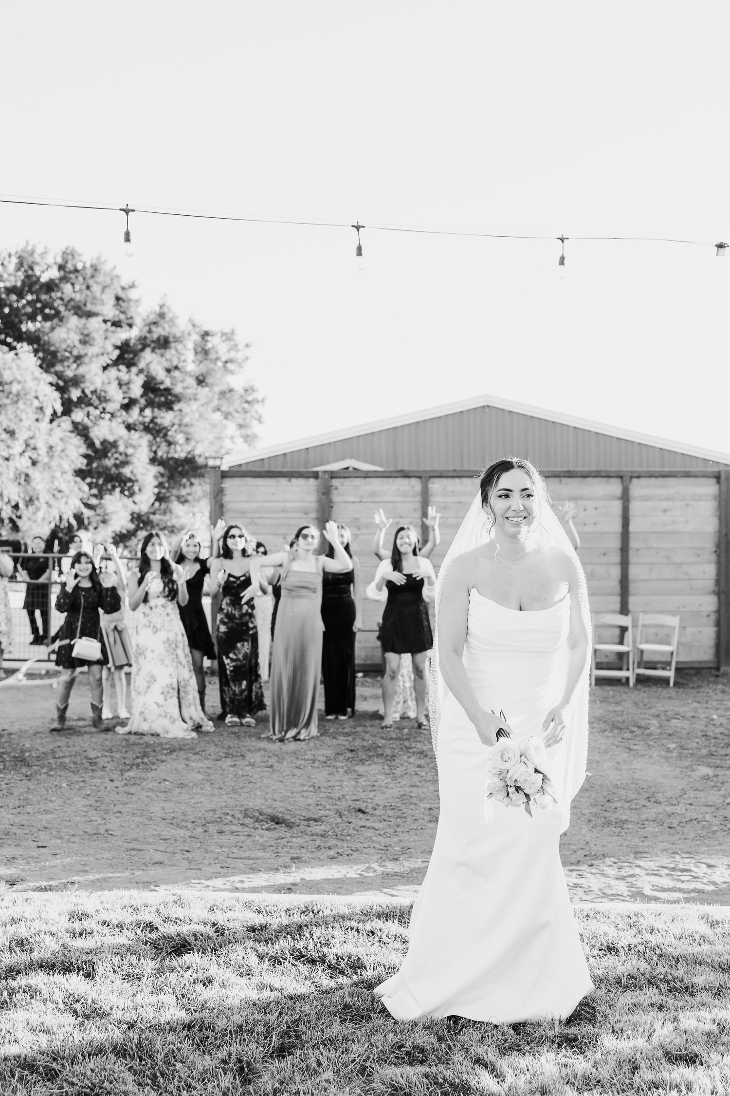 Bride in a white wedding gown poses in a sunlit field with guests in the background.