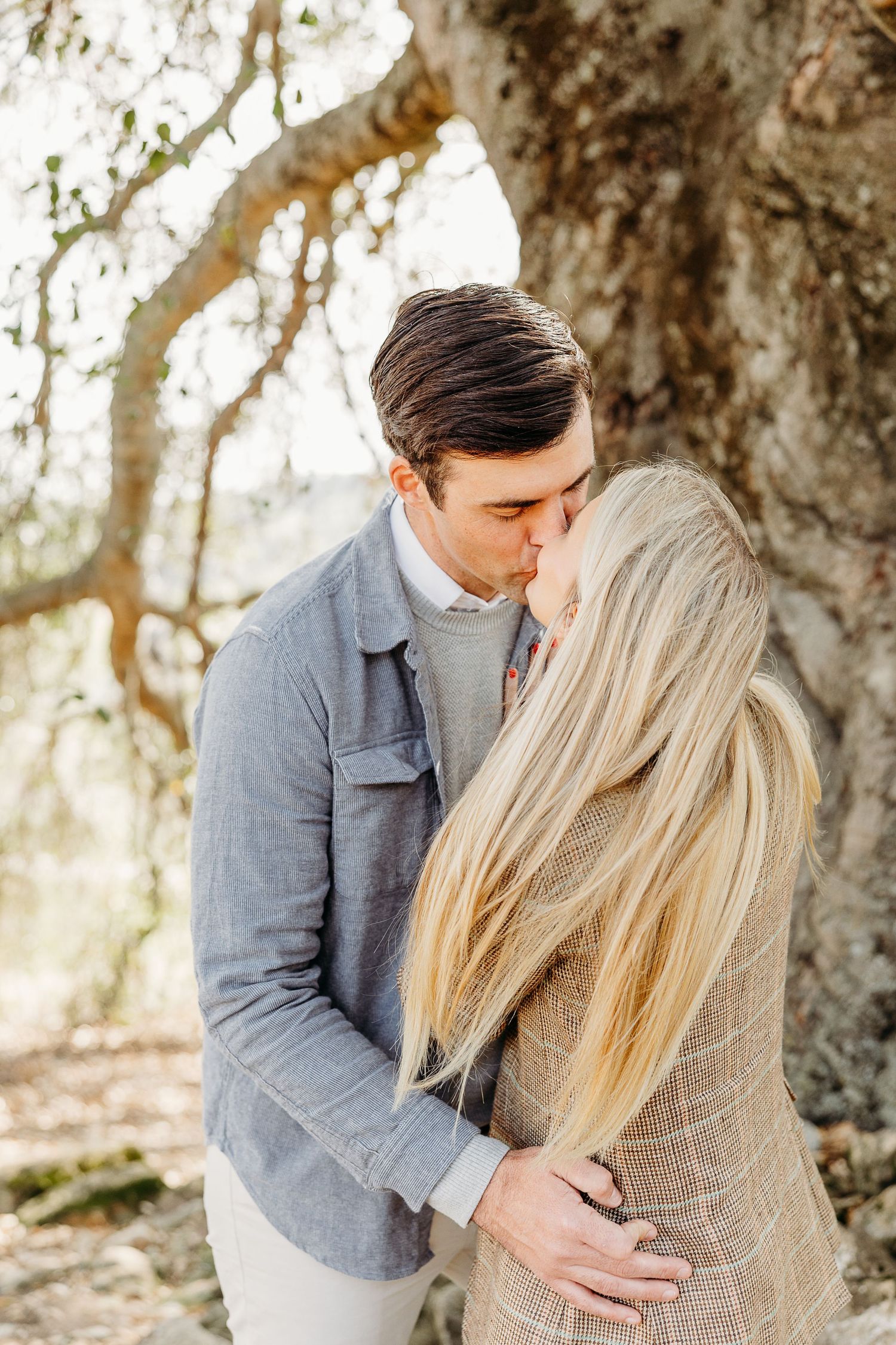A romantic couple embraces in a sunlit forest setting wearing coordinated grey and neutral toned outfits.