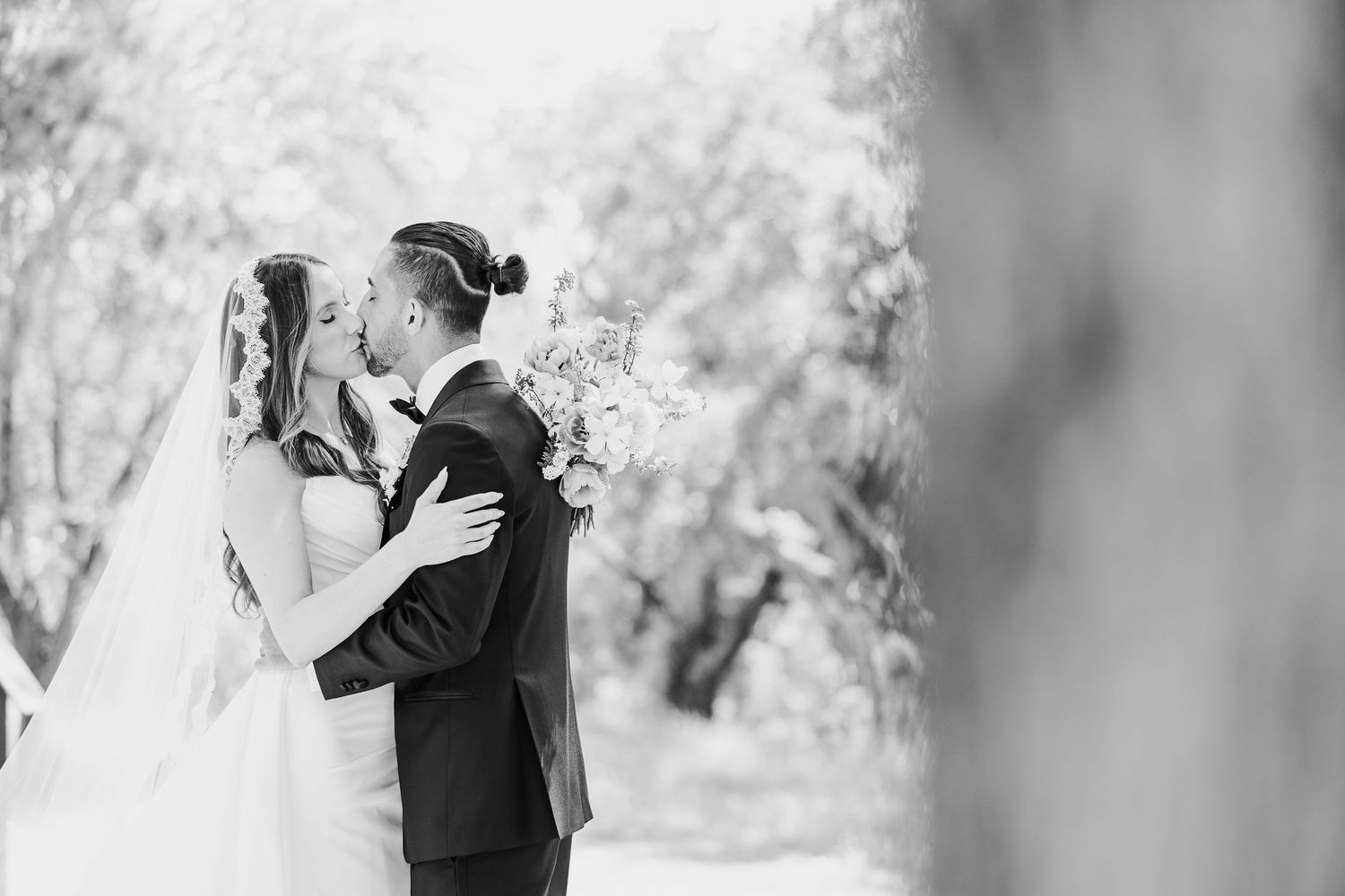 Black and white wedding photo of bride and groom sharing a romantic moment outdoors among blossoming trees.