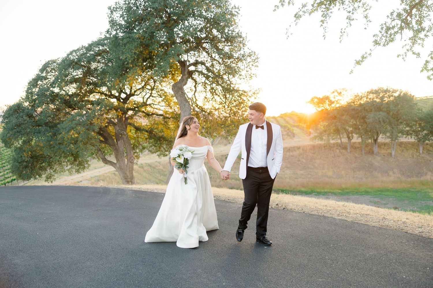 Bride and groom walk hand-in-hand at sunset on a scenic path, surrounded by golden trees at their outdoor wedding.