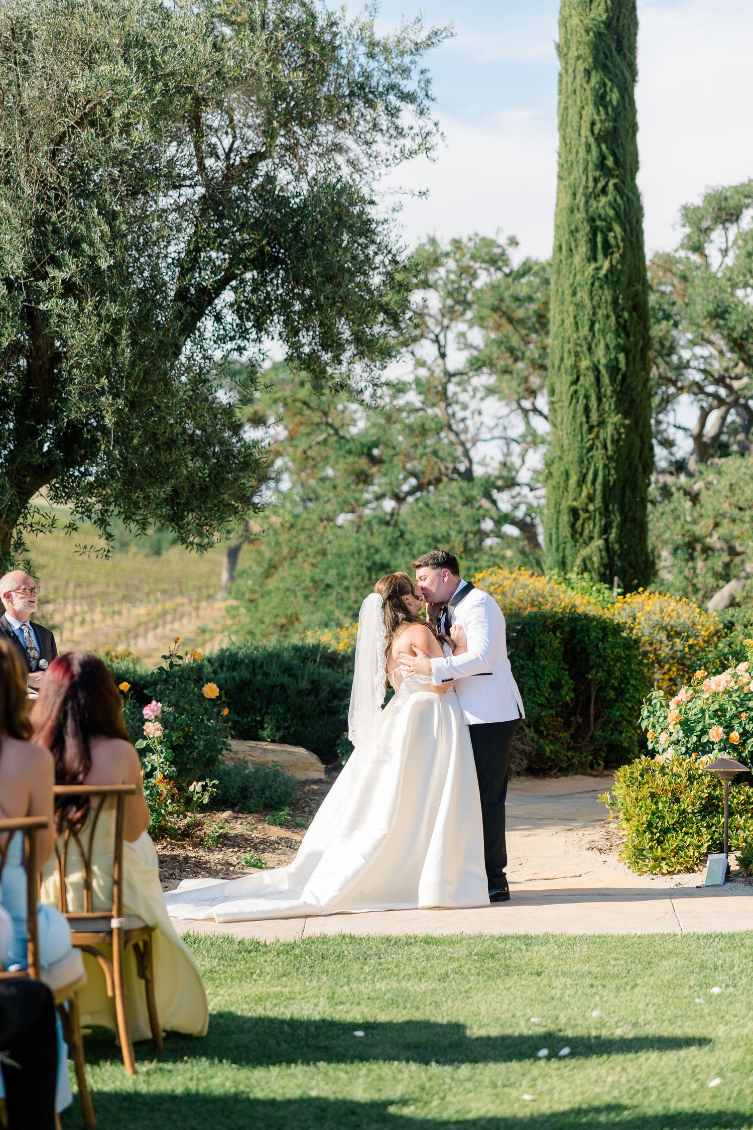 Bride and groom share first kiss at outdoor garden wedding ceremony with cypress trees.