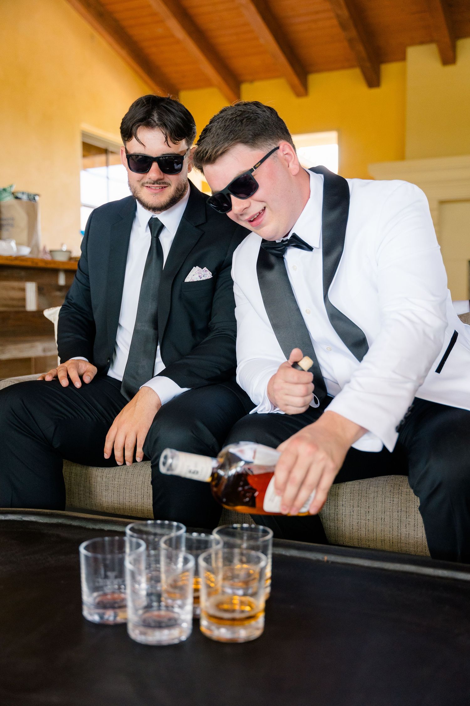 Groom and groomsman in tuxedos with sunglasses pouring whiskey before the wedding ceremony.