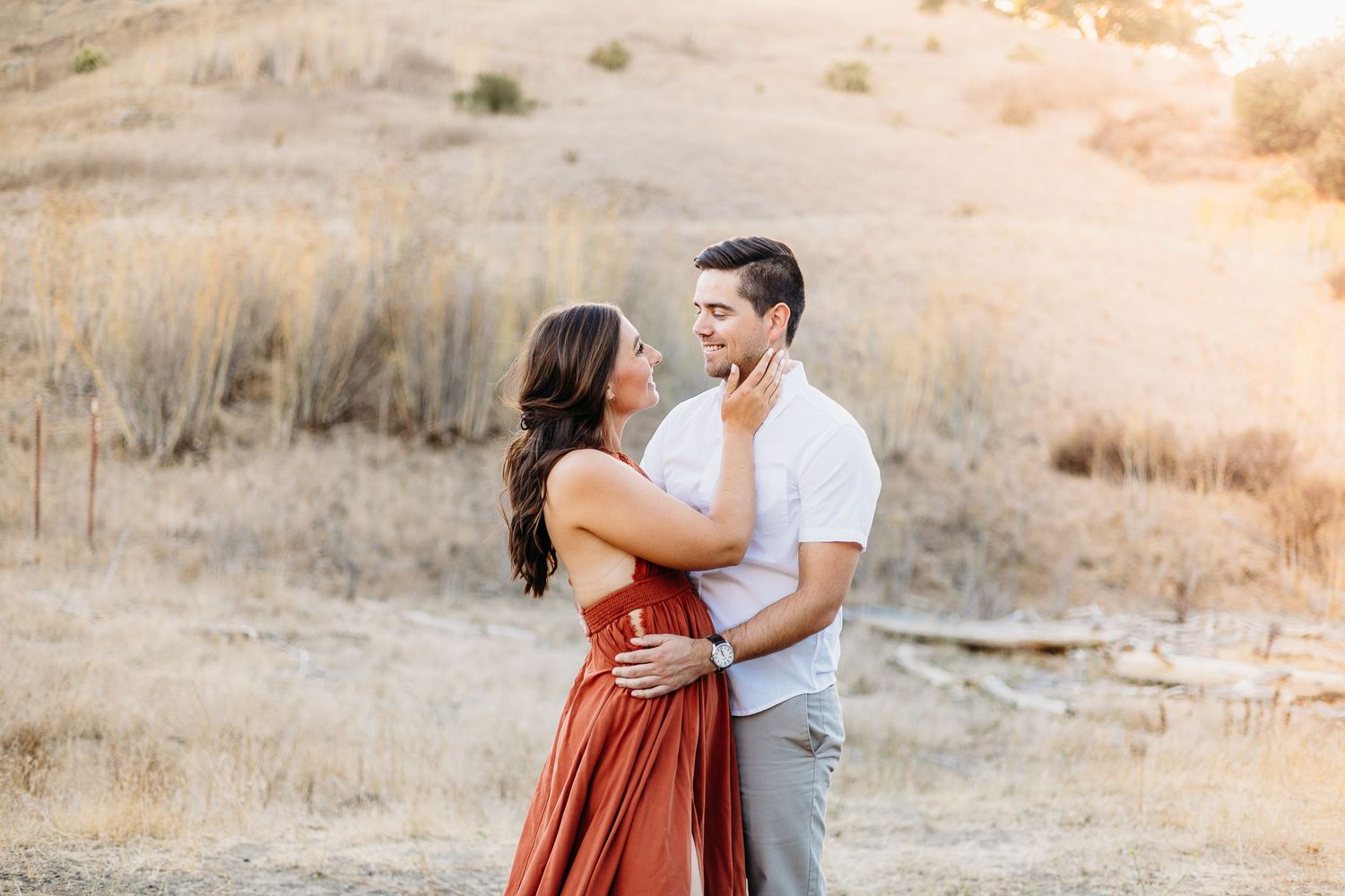 A romantic couple embracing in golden sunlit field wearing rust colored dress and white shirt.