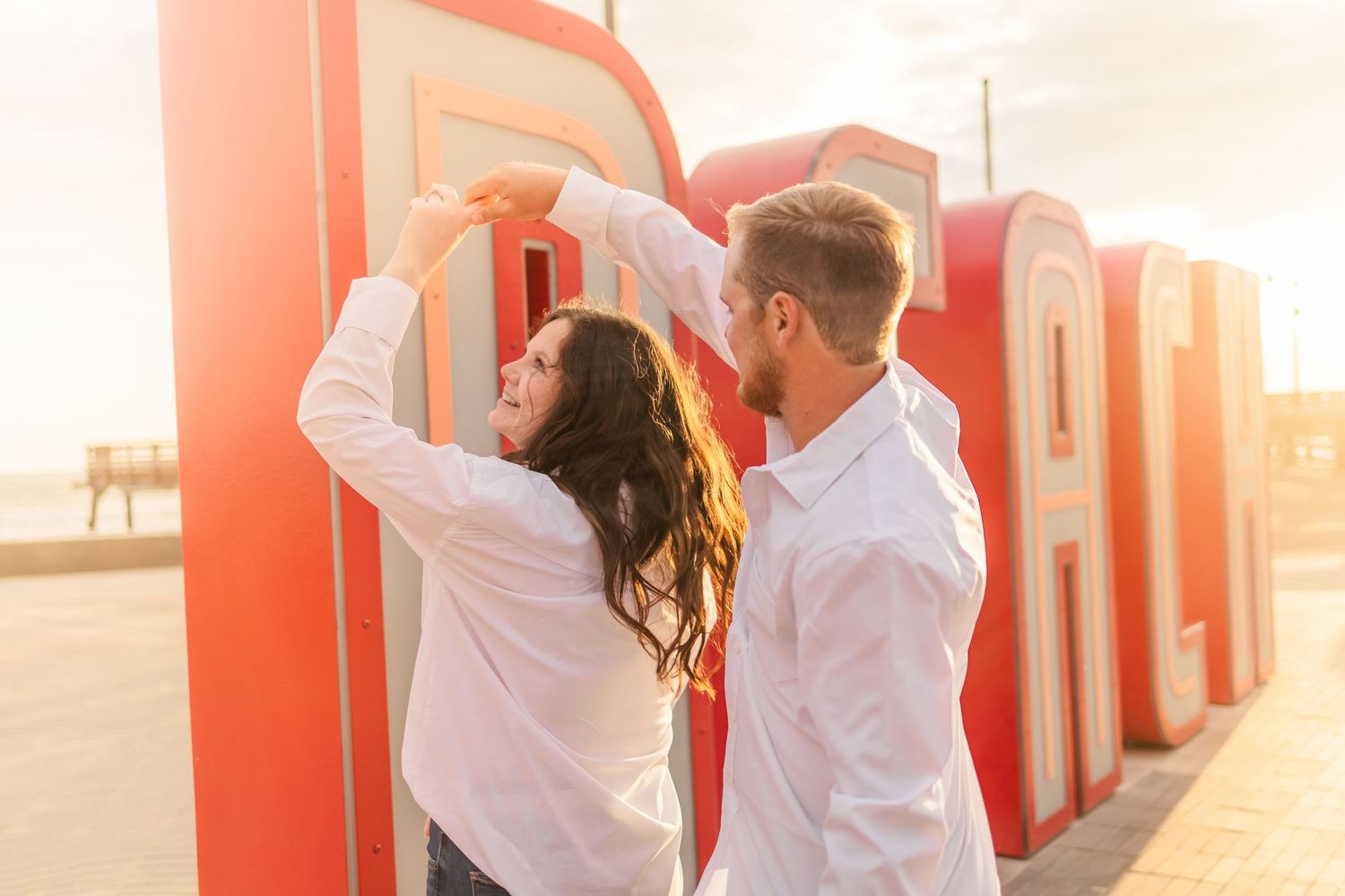 Dancing couple in matching white shirts poses playfully in front of red outdoor lettering at sunset.