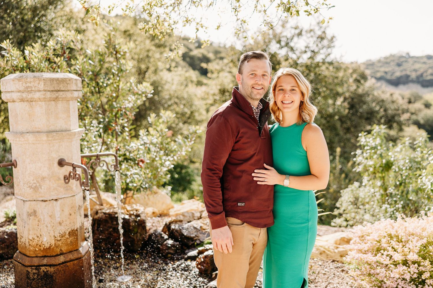 A couple shares a romantic moment outdoors among blooming spring trees in a pastoral mountain setting.