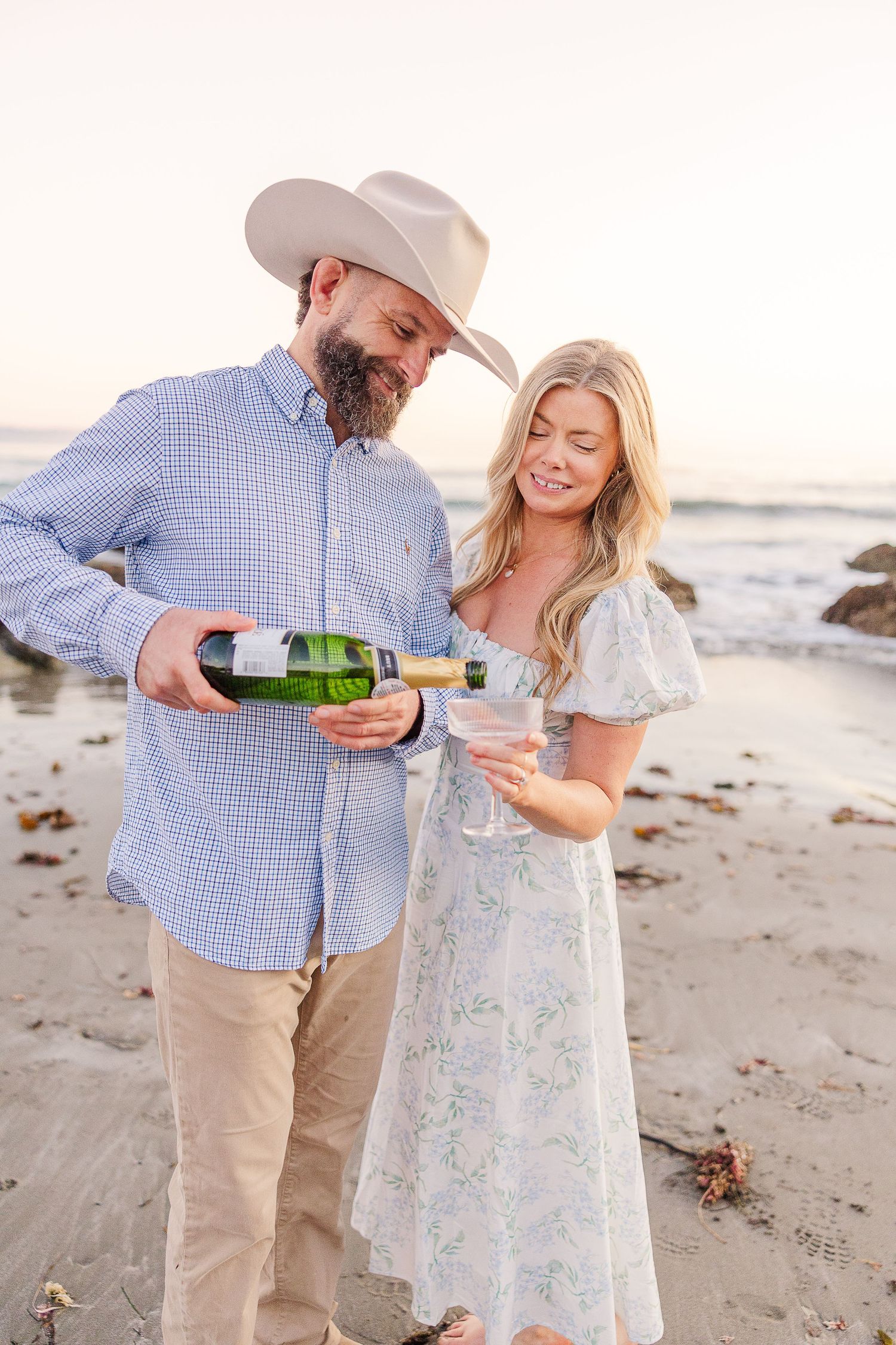 Couple celebrating with champagne on beach during golden hour engagement photoshoot.