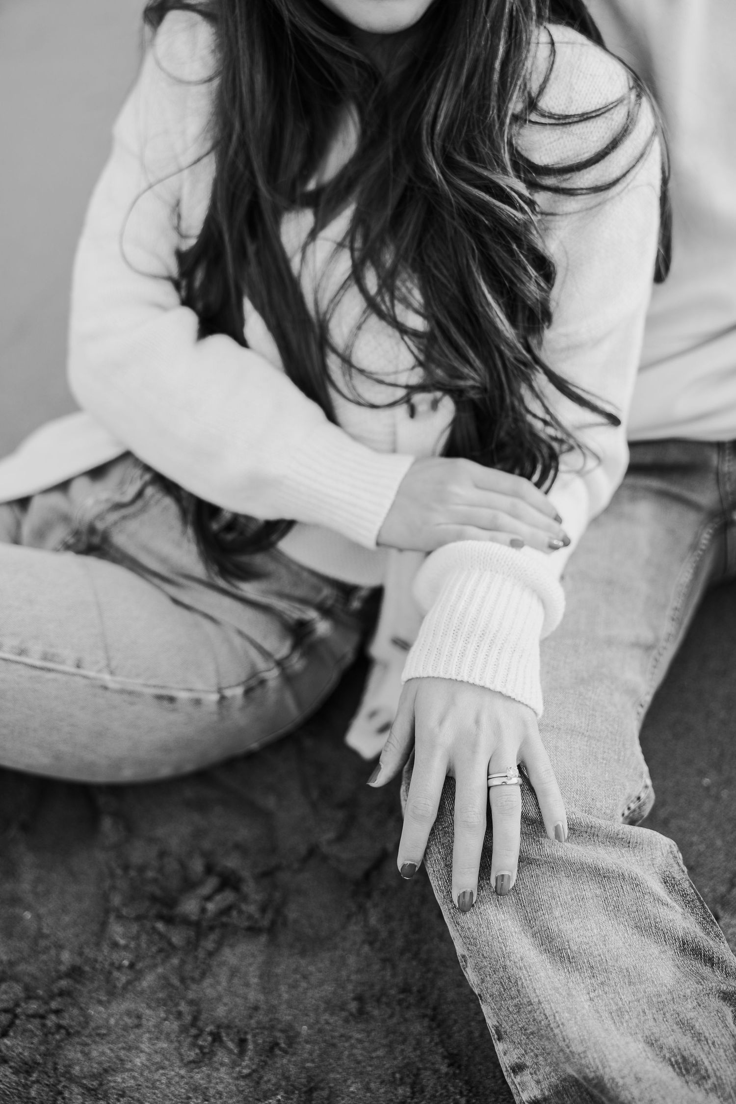 Black and white photo showing person in white sweater and jeans sitting casually with hand extended displaying ring.