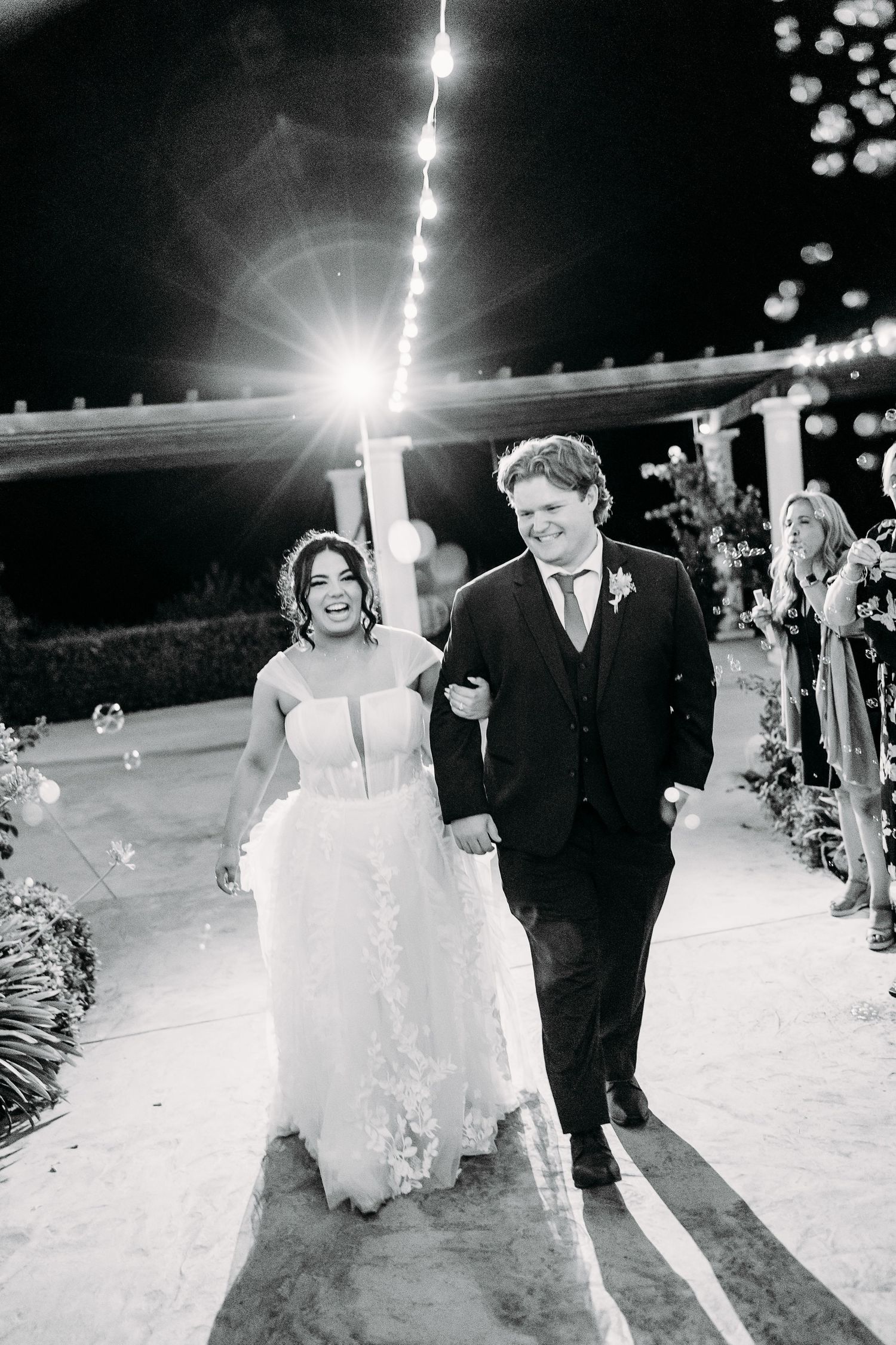 Newlyweds walking together under romantic string lights at nighttime outdoor venue.