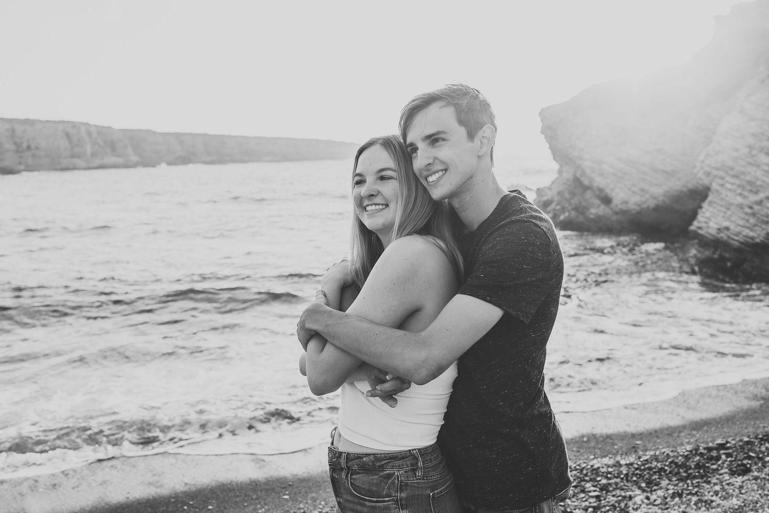 A black and white photo of a couple hugging on a beach with coastal cliffs behind them.