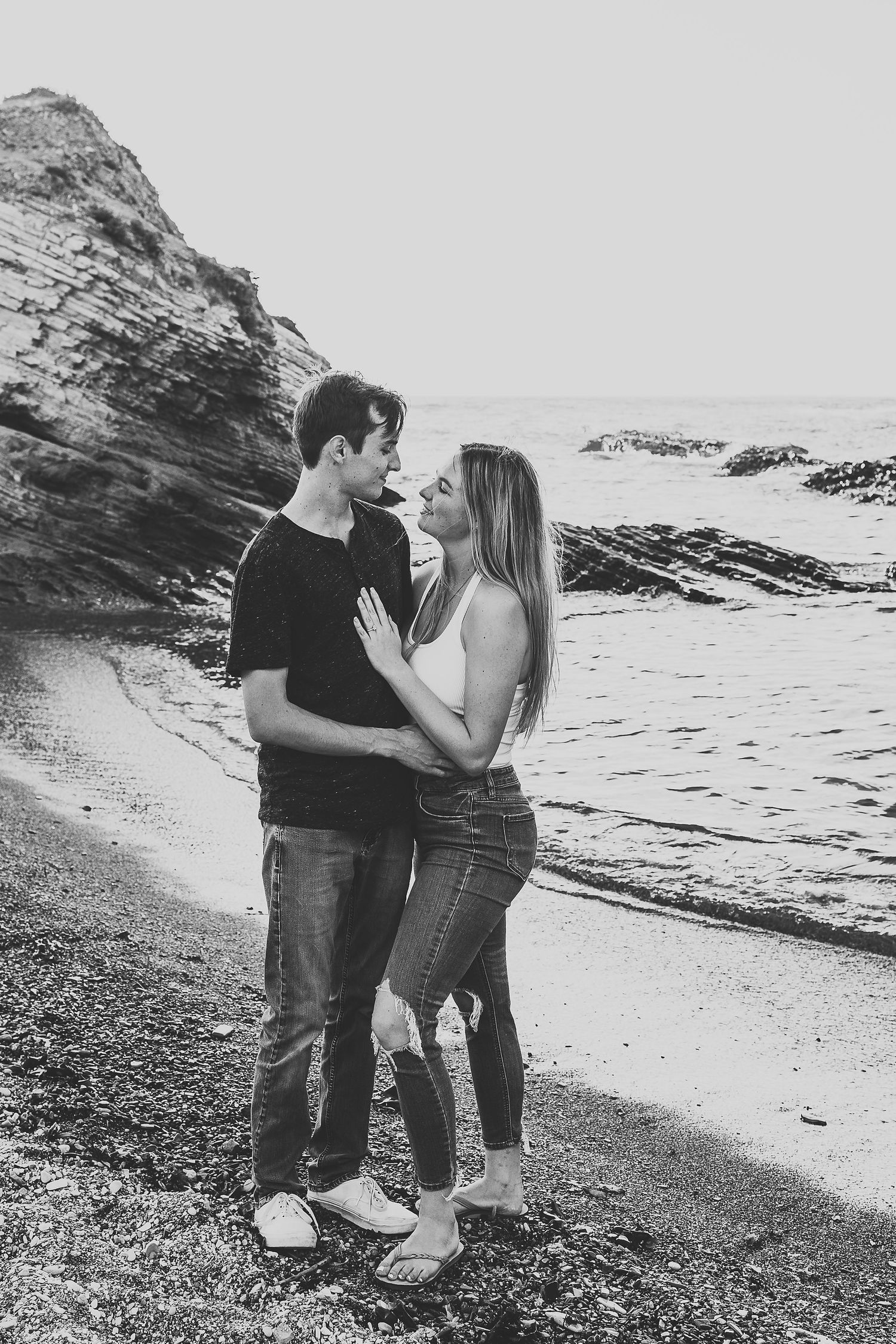 A black and white photo of a couple embracing on a rocky beach with cliffs in the background.