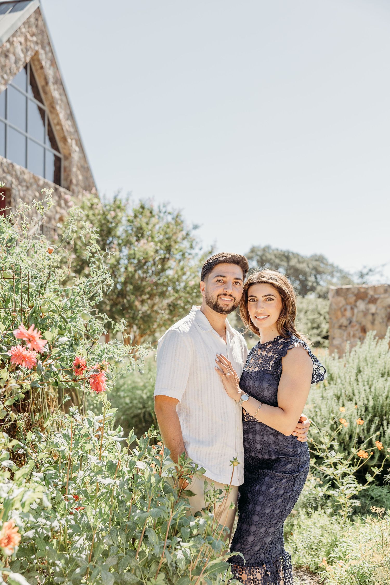 Engagement photo in garden with rustic building and blooming flowers.