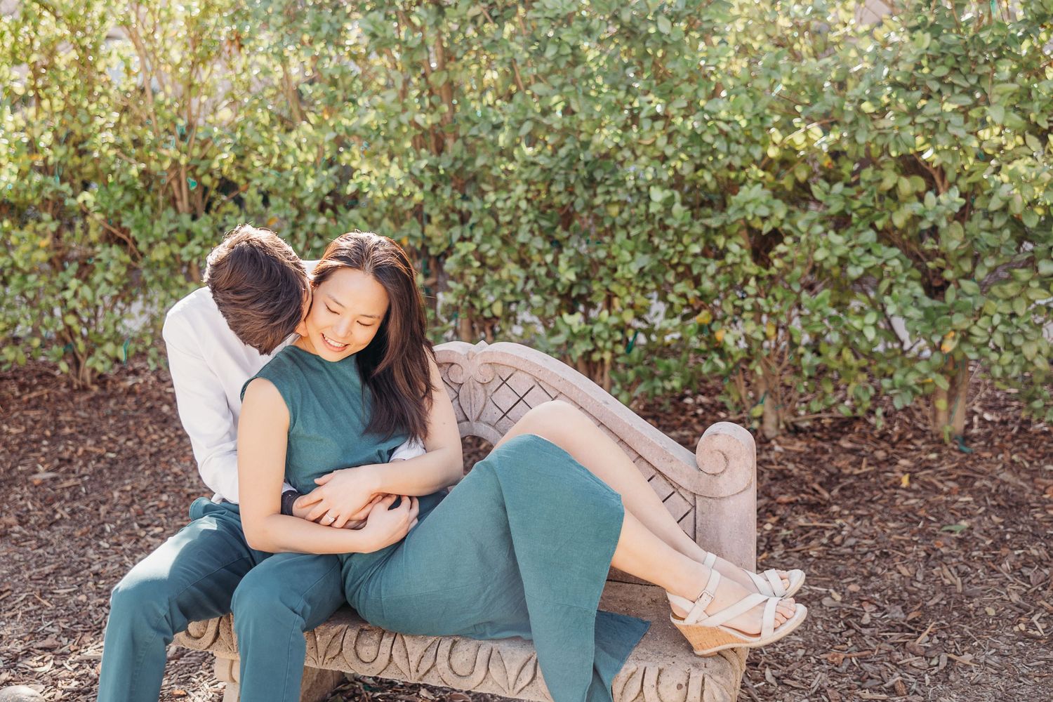 Couple relaxes together on outdoor stone bench against green foliage background.