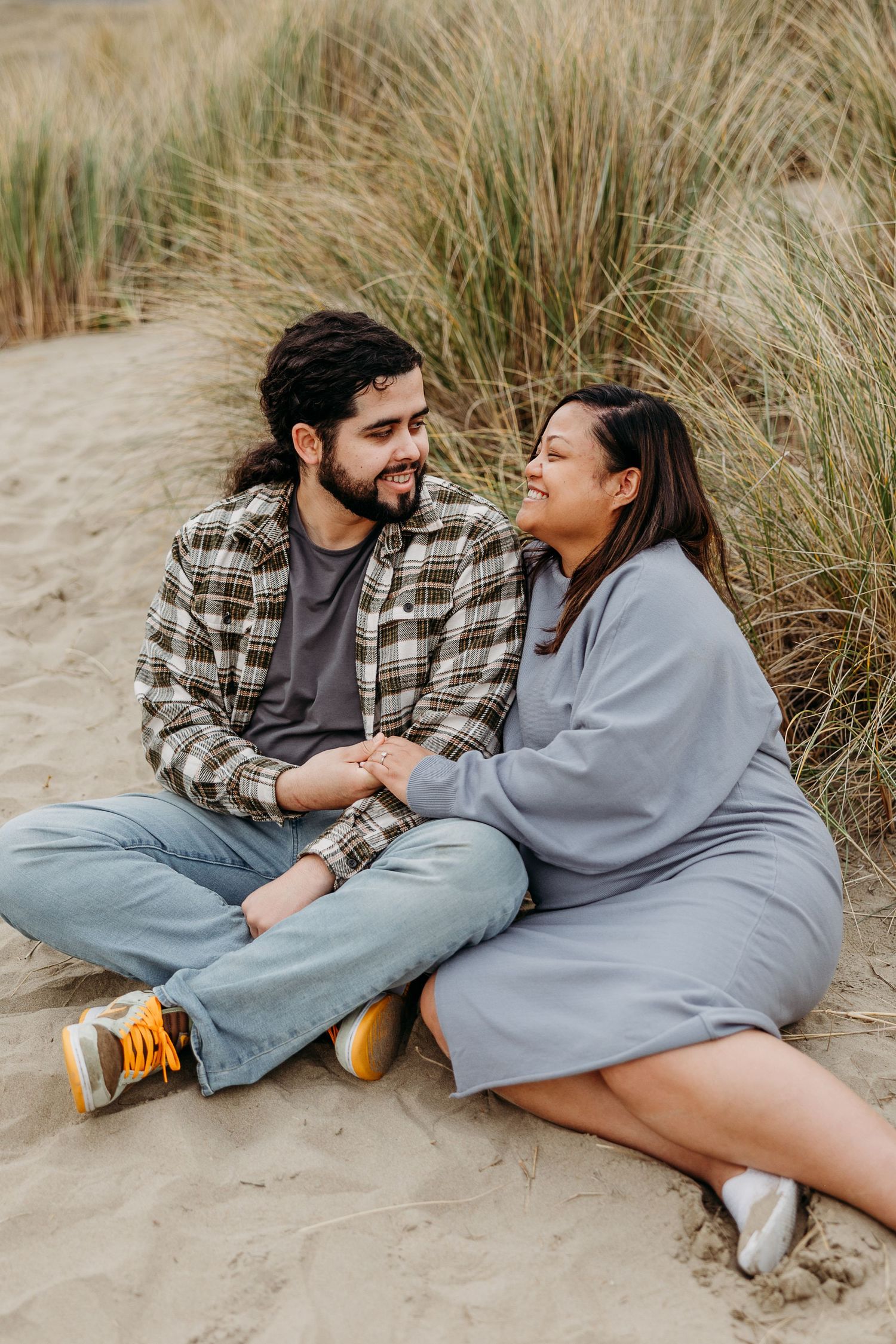 A couple sitting together on sandy dunes with beach grass, sharing an intimate moment by the coast.