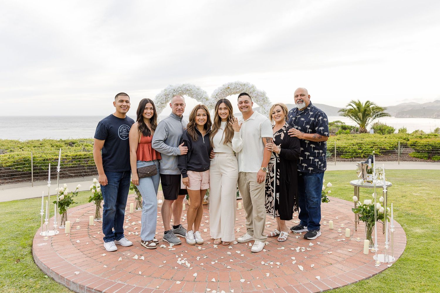 Group of people posing together on a grassy lawn near a beach with palm trees in the background.