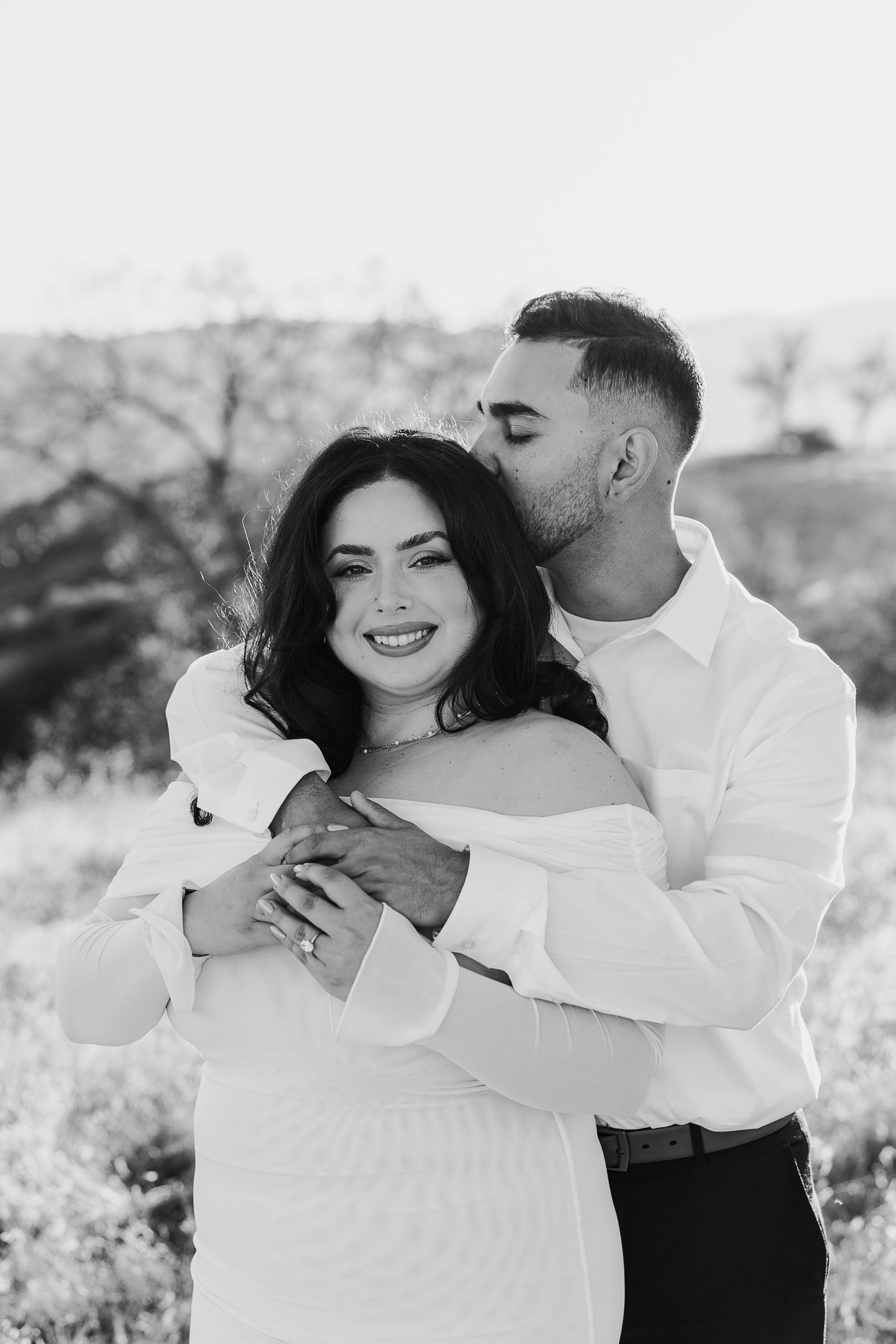 A romantic black and white portrait of a couple embracing outdoors in white attire.