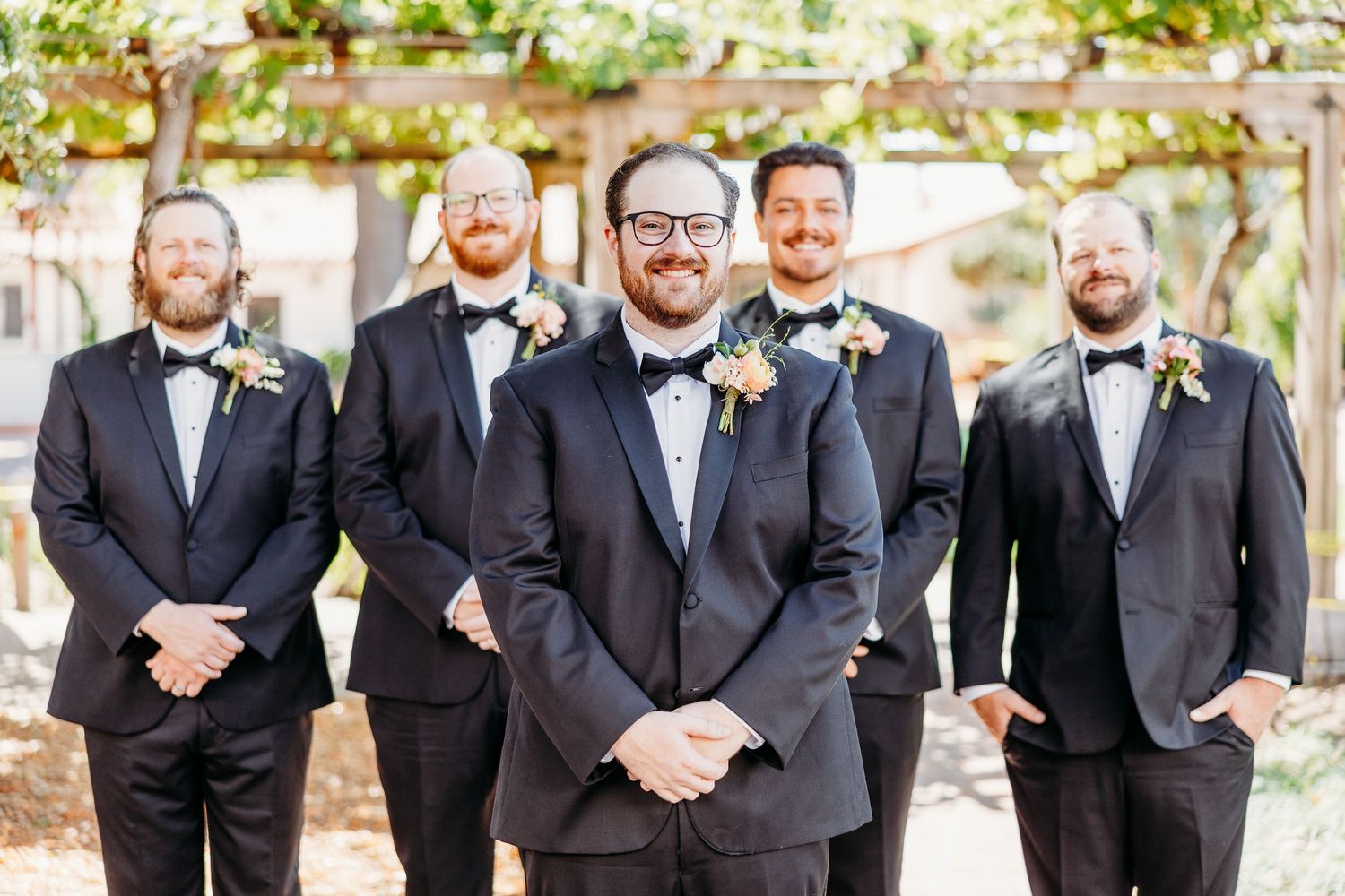 Groomsmen in matching black tuxedos pose under garden pergola during wedding celebration.