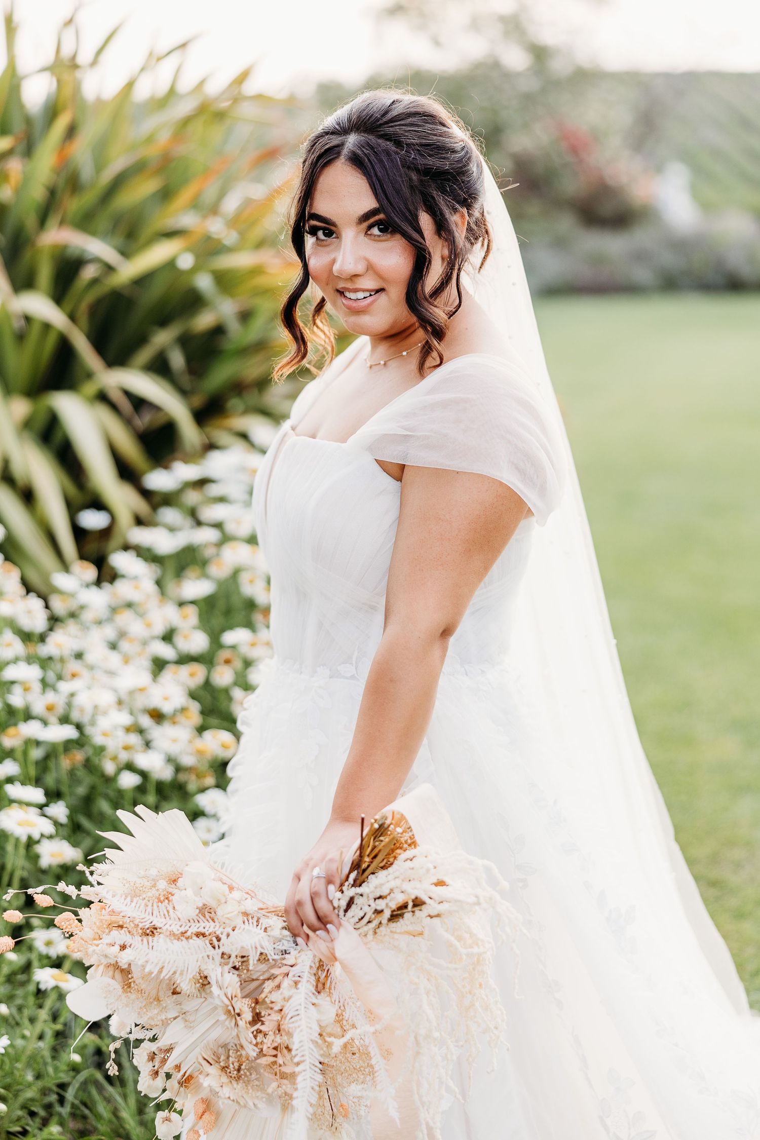 Bride in white cap sleeve wedding dress holding dried flower bouquet poses in garden setting.