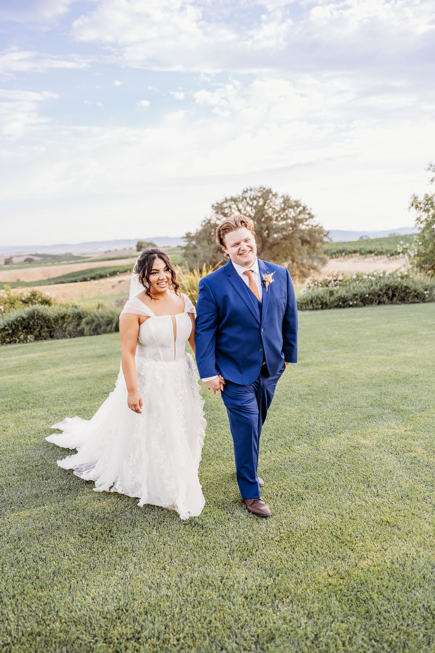Wedding couple walking together on lush green lawn with scenic countryside backdrop.