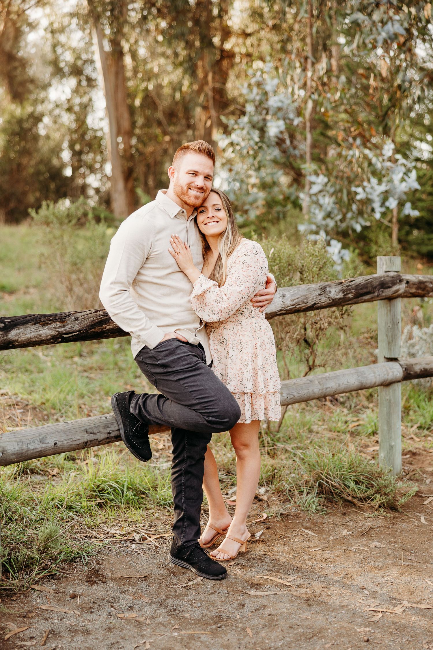 Couple posing playfully by wooden fence in woodland setting during sunset.